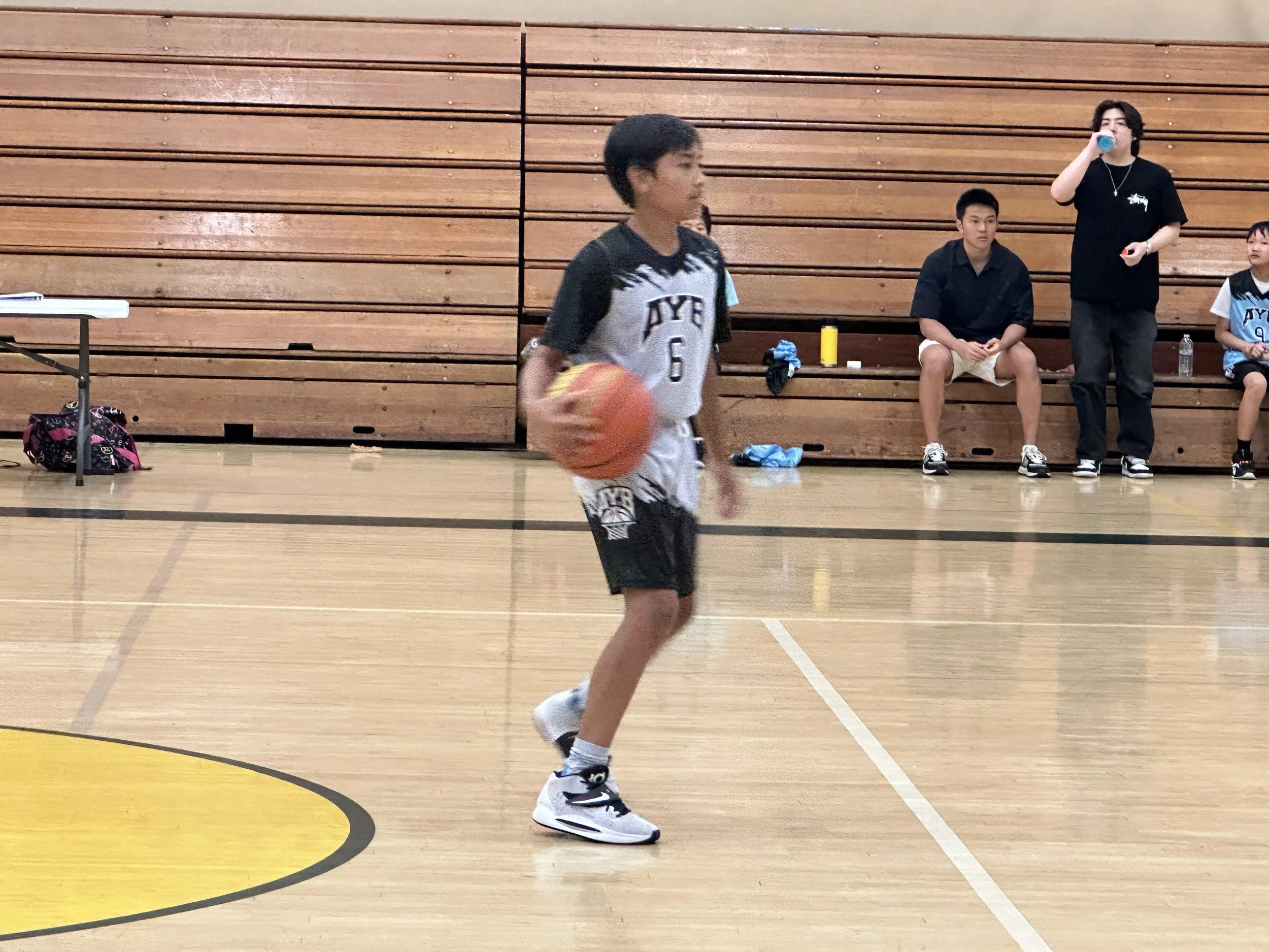 Young basketball player dribbling on a gym court with spectators in background.