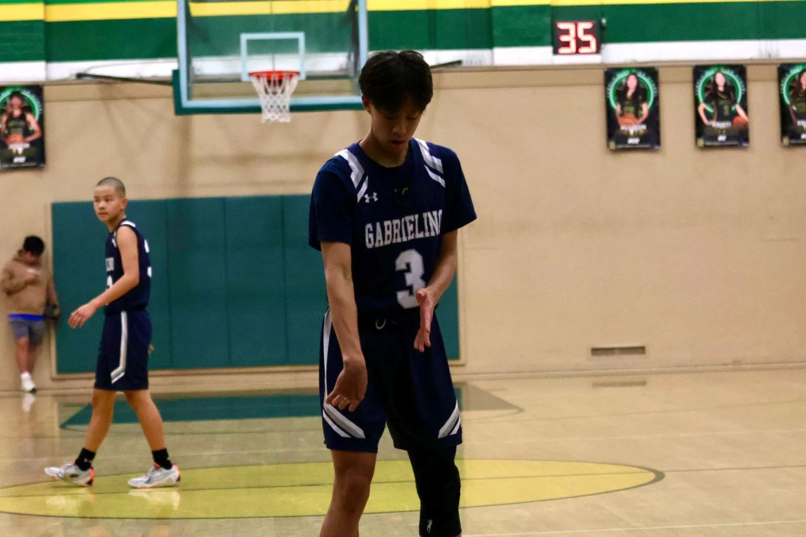 High school basketball players on a court during a game with jerseys featuring the word "Gabrielino." A scoreboard is visible in the background.