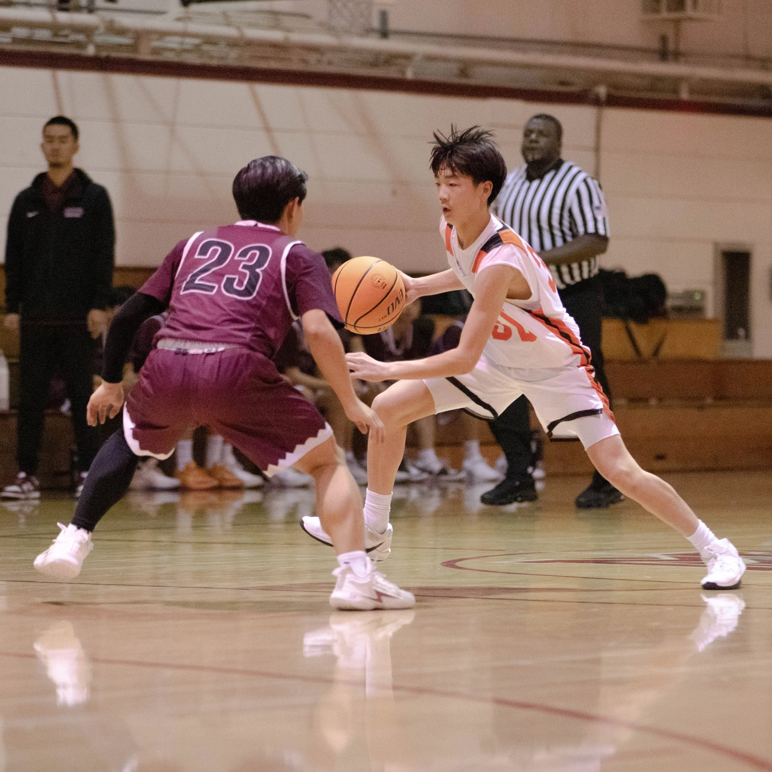 Two high school basketball players in action during a game indoors, one dribbling while facing a defender. A referee and another person are visible in the background.