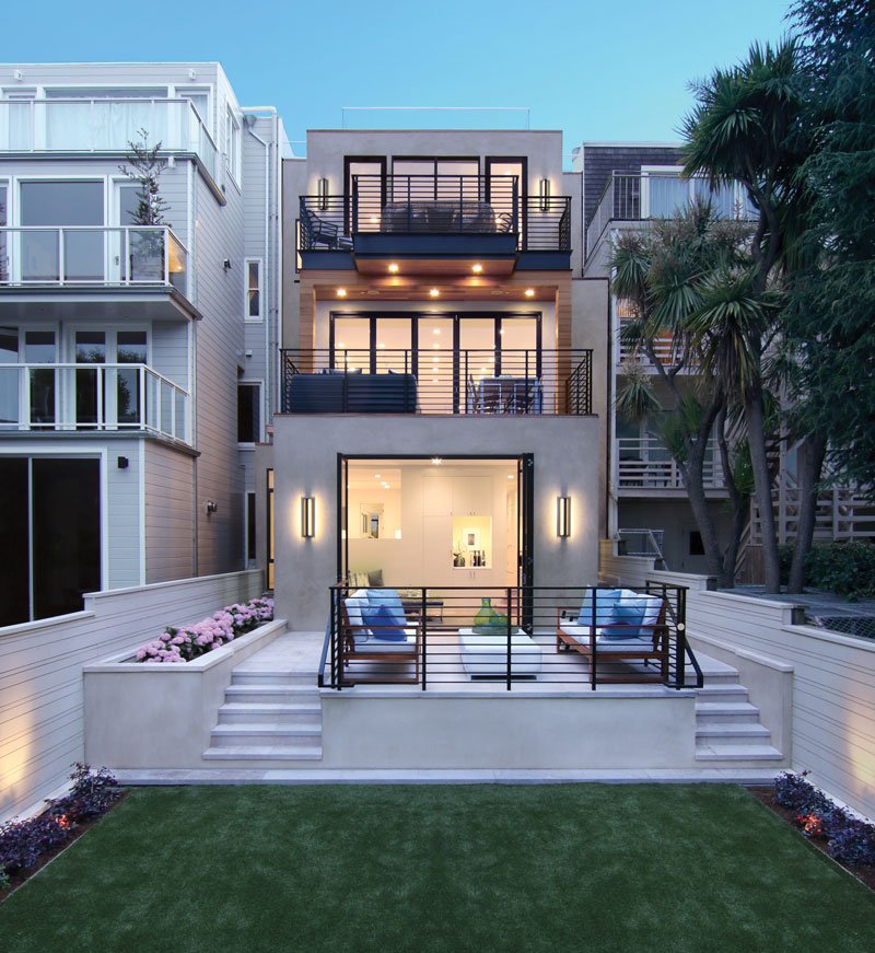 Modern three-story house with balconies and patio, large windows, and a well-manicured lawn, surrounded by neighboring homes under a clear blue sky.