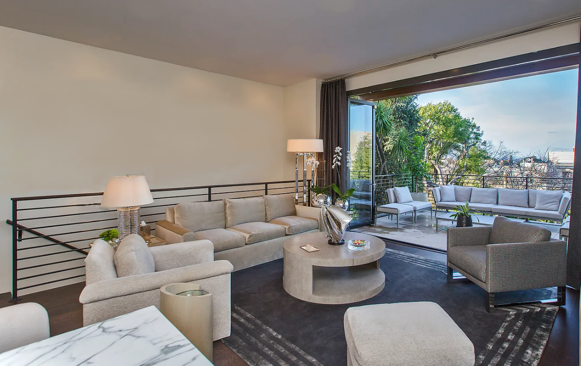 Modern living room with beige sofa, armchairs, coffee table, decorative plants, and large sliding glass doors opening to a balcony.