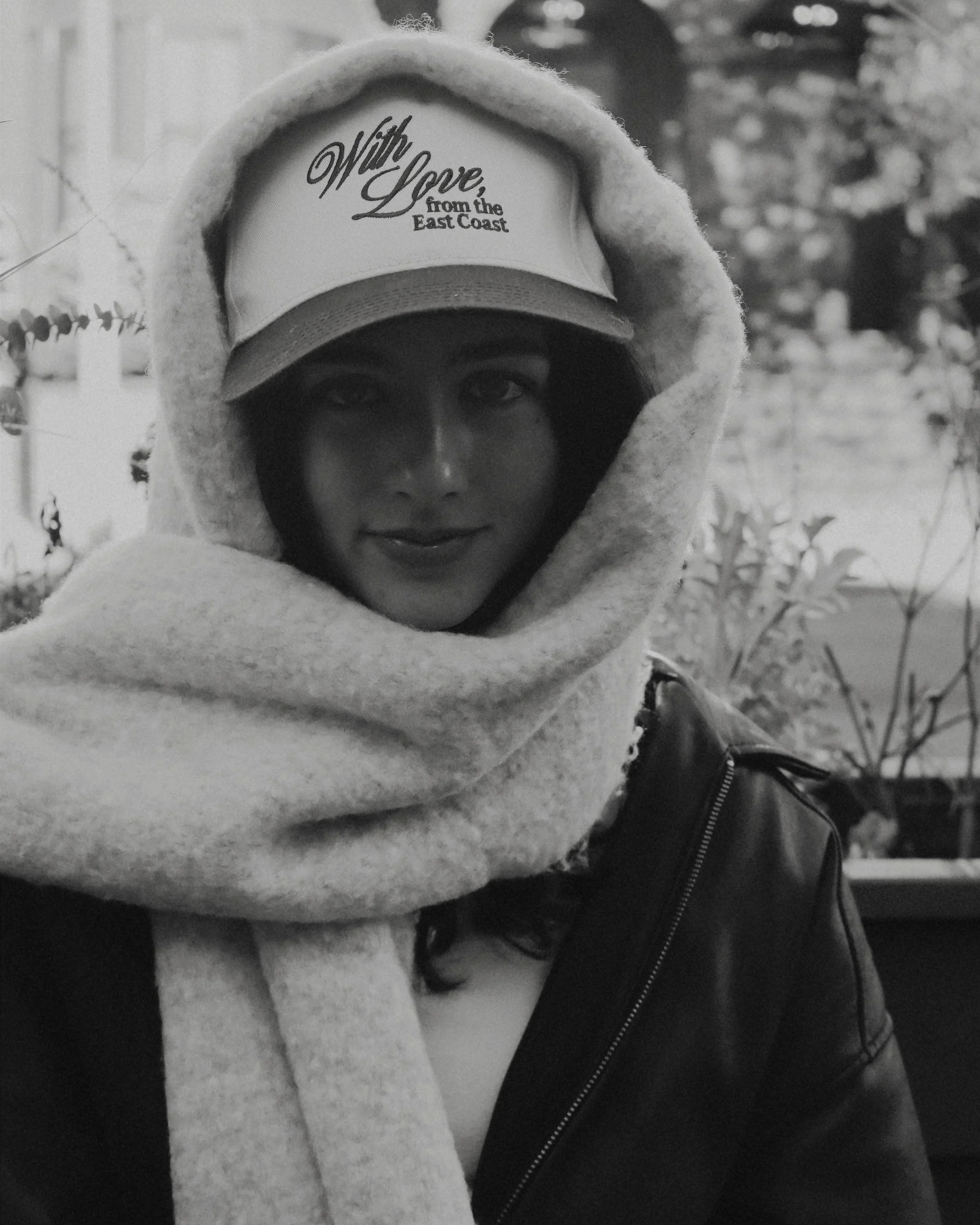 A woman wearing a cap that reads 'With Love from the East Coast,' a hooded scarf, and a leather jacket.