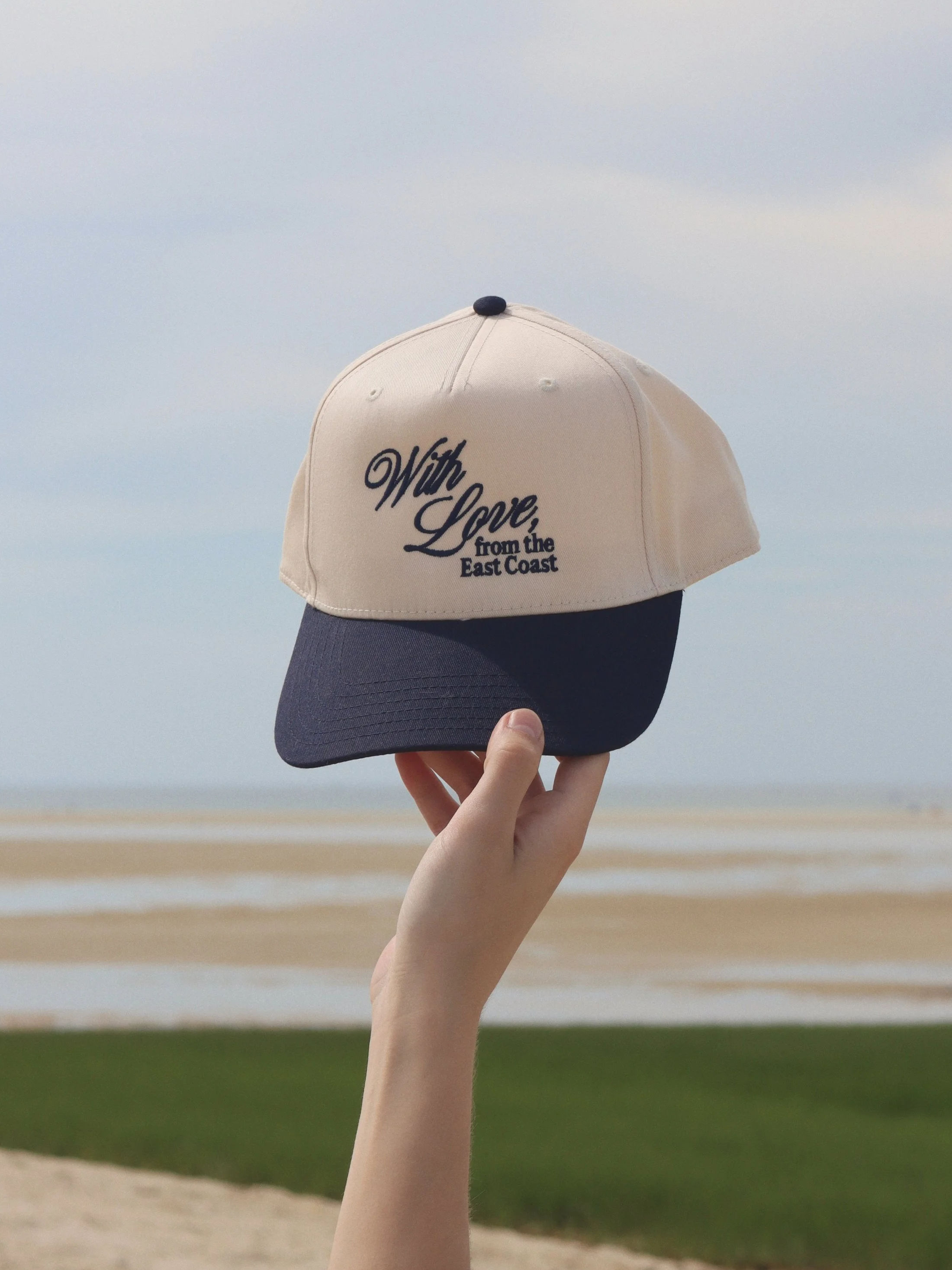 Hand holding a beige and navy baseball cap with the words "With Love, from the East Coast" embroidered on the front, against a background of a beach with water and sky.