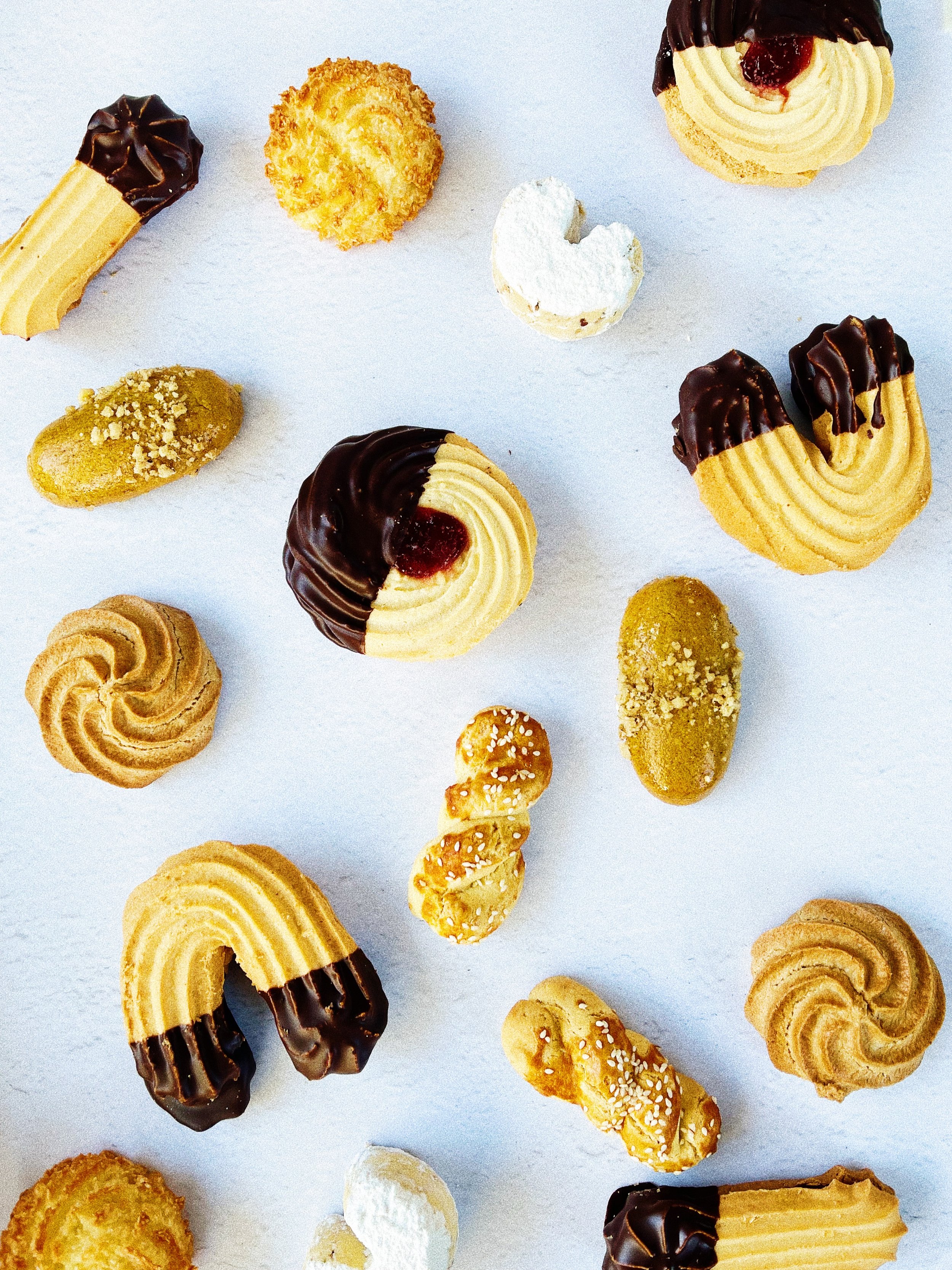 Assorted Greek cookies and biscuits on a white background, including choc dipped shortbread, coconut macarons, cookies with chocolate and cream, and biscotti.