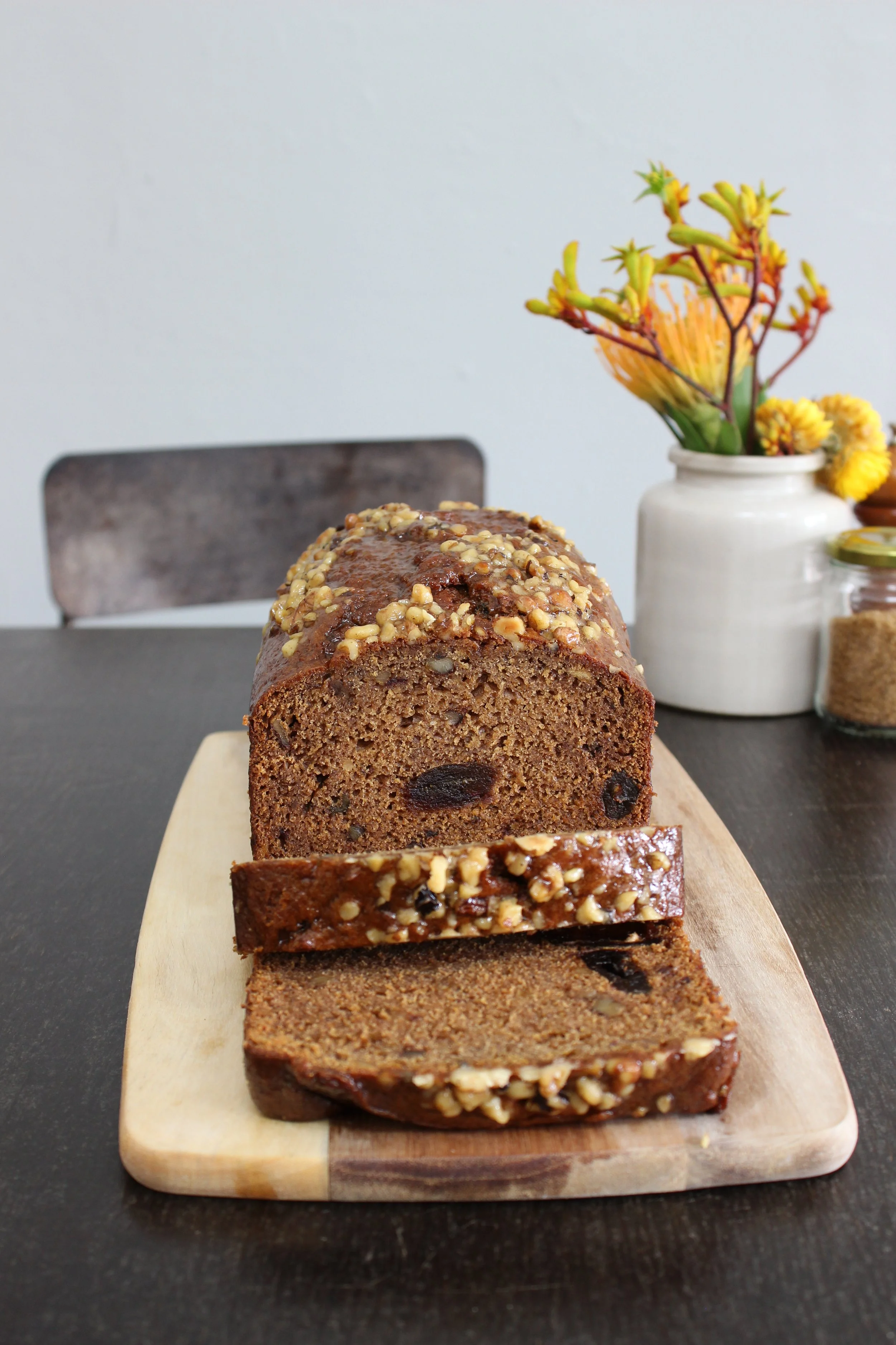 A fruit & nut loaf cake with chopped nuts on top, partially sliced on a wooden serving board, on a dark table with a vase of colorful flowers in the background.