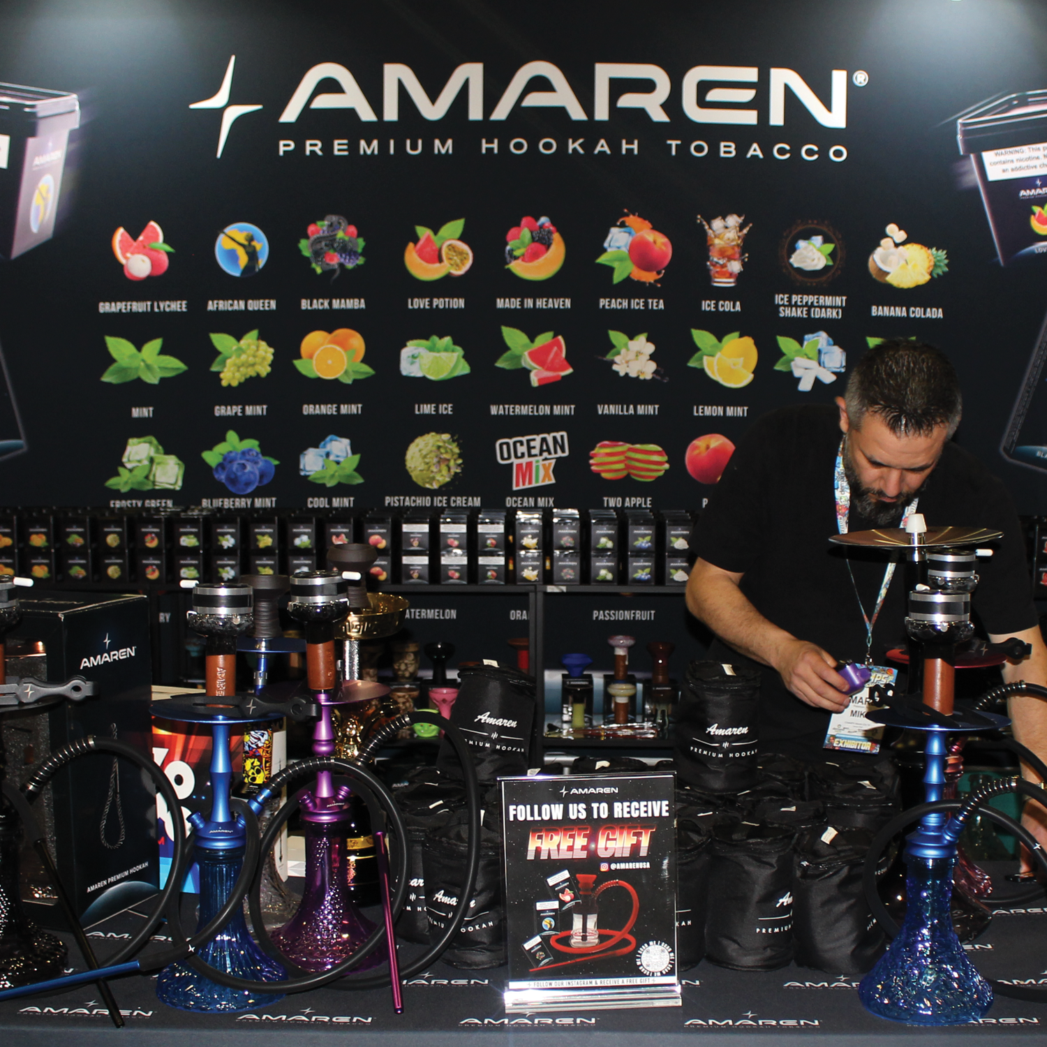 Man setting up hookah pipes at Amaren Premium Hookah Tobacco booth with a display of various tobacco flavors in the background.