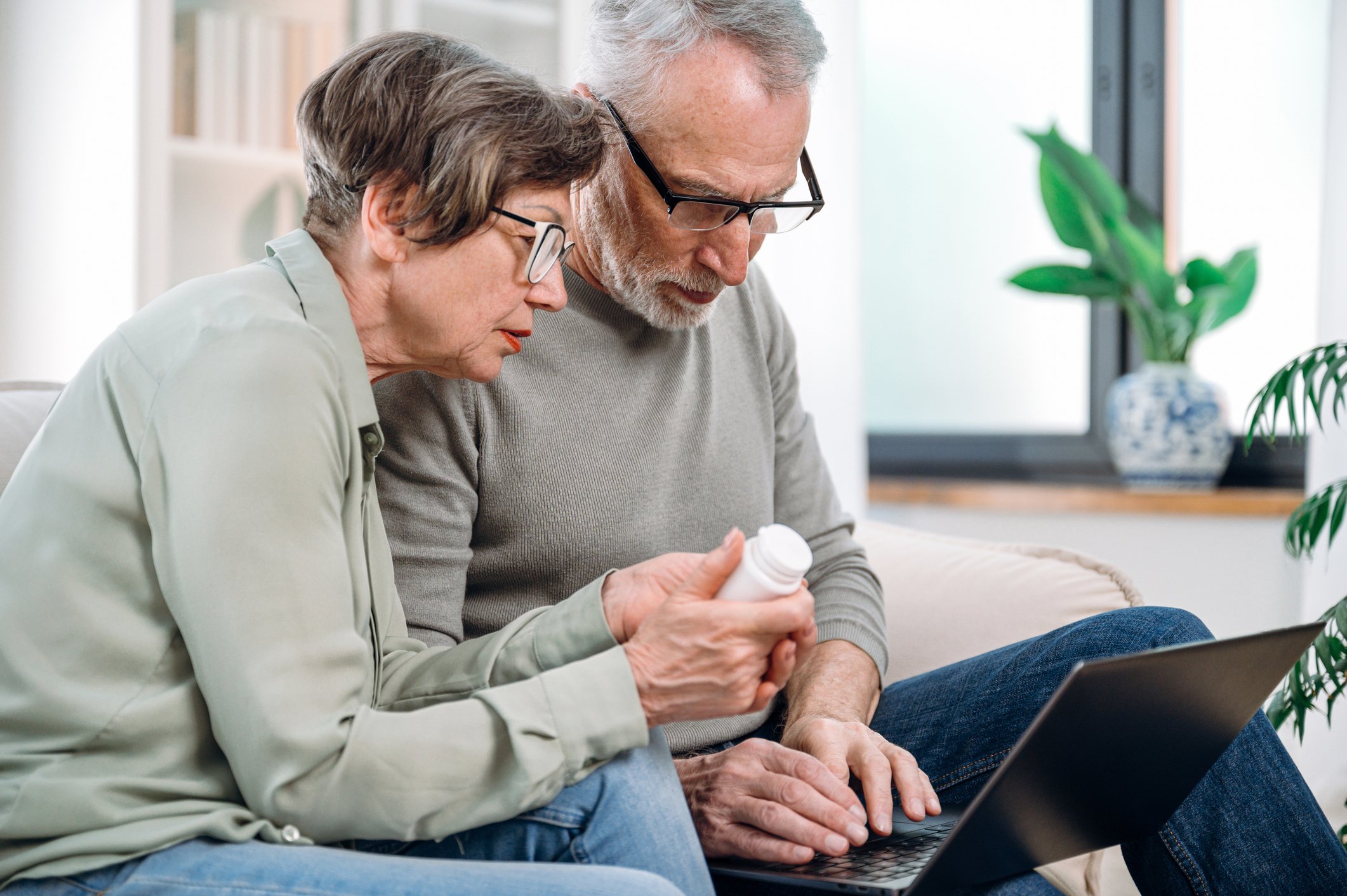 An elderly man and woman sitting closely together on a beige couch, looking at a tablet and sharing a prescription bottle, in a bright room with a window and a green potted plant.