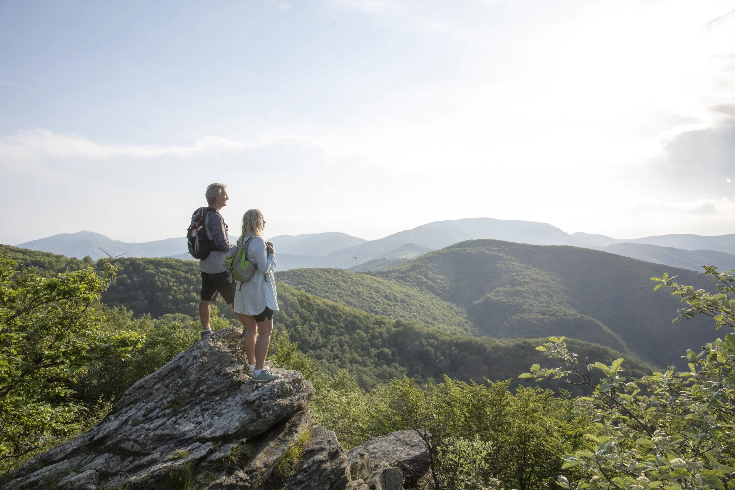 Two hikers, one man and one woman, standing on a large rock overlooking a lush green mountain landscape under a bright sky.
