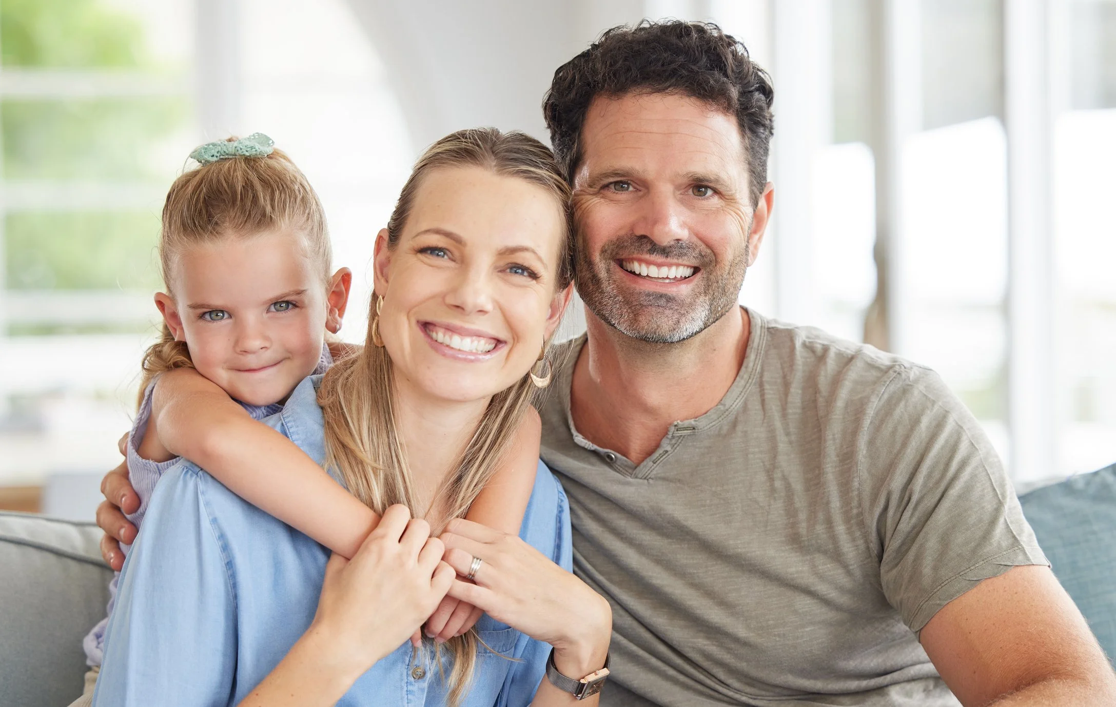 A happy family of three sitting on a couch in a bright living room, with the mother and father smiling and the daughter hugging her mother from behind.