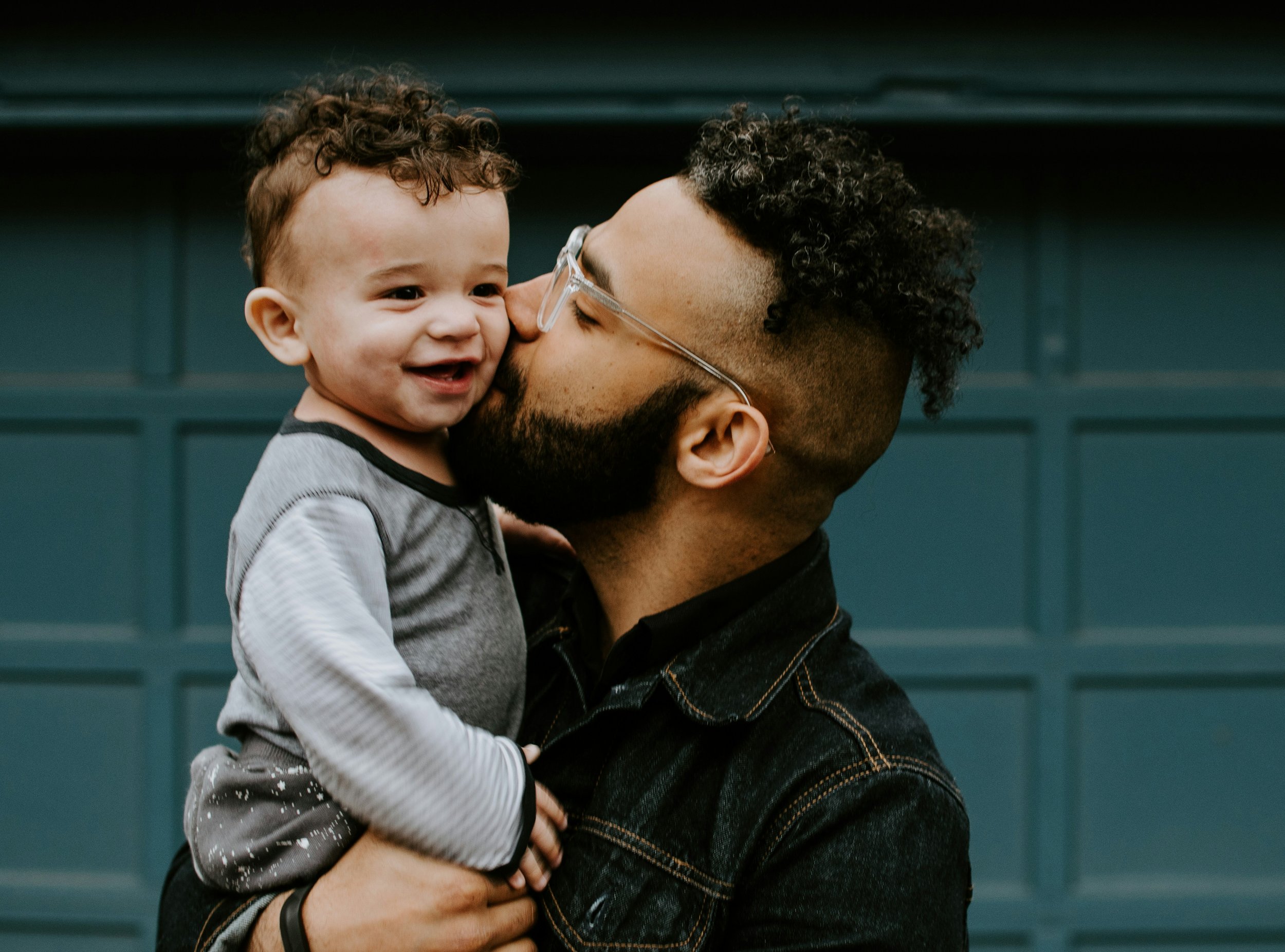 A man with glasses and a beard holding a smiling young boy with curly hair and a gray and black striped shirt. They are close to each other, sharing a tender moment in front of a teal wall.