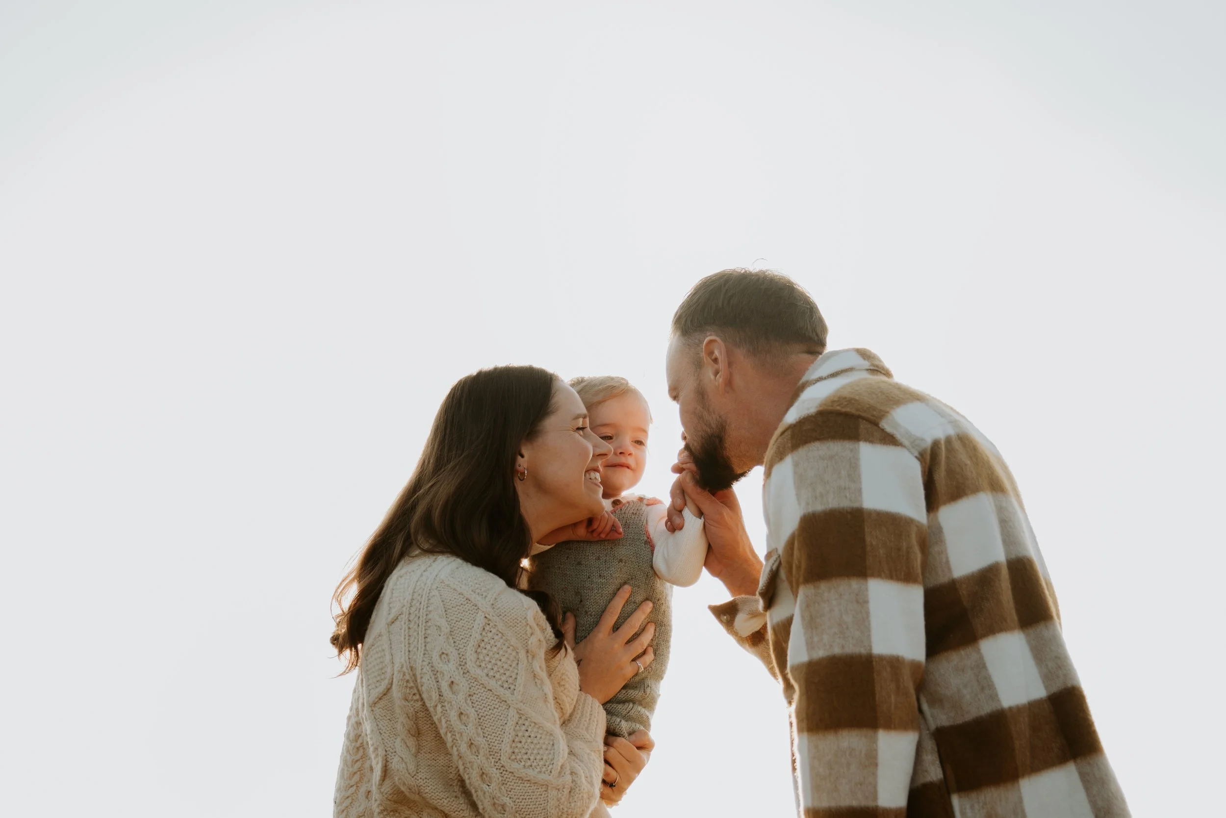 Mother holding child and father kissing childs hand backlit at sunset photographed by an Ocean Grove family photographer