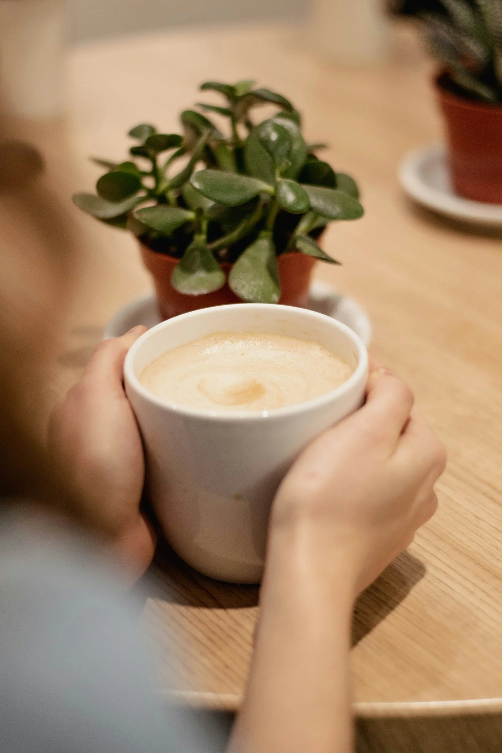 Open hands holding a white mug of warm comforting liquit on a wooden table, with a plant in the background, ready to discuss grief.