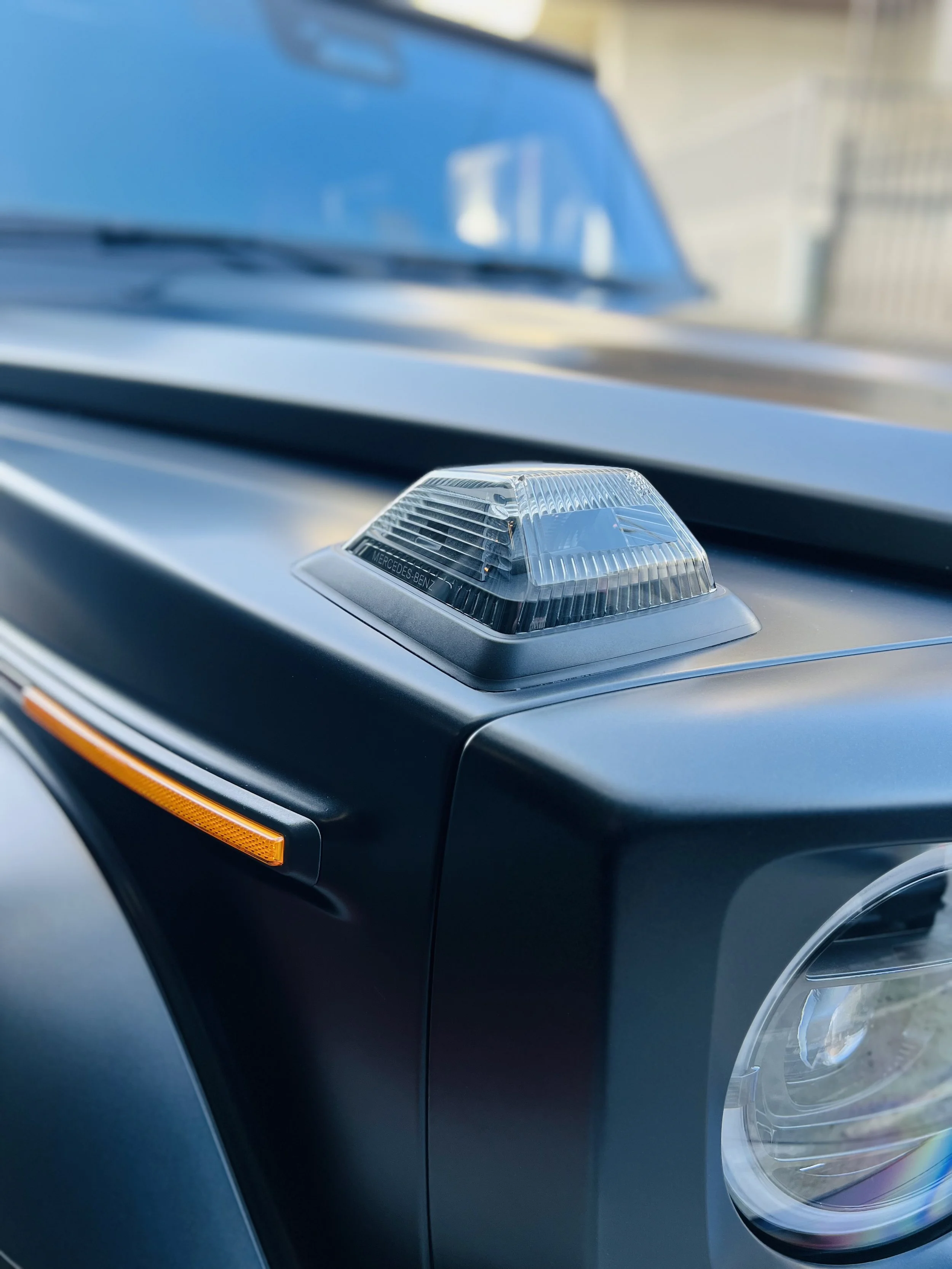 Close-up of a black vehicle's front with a clear round headlight and an amber side marker light, featuring a rooftop emergency light with the Mercedes-Benz logo.