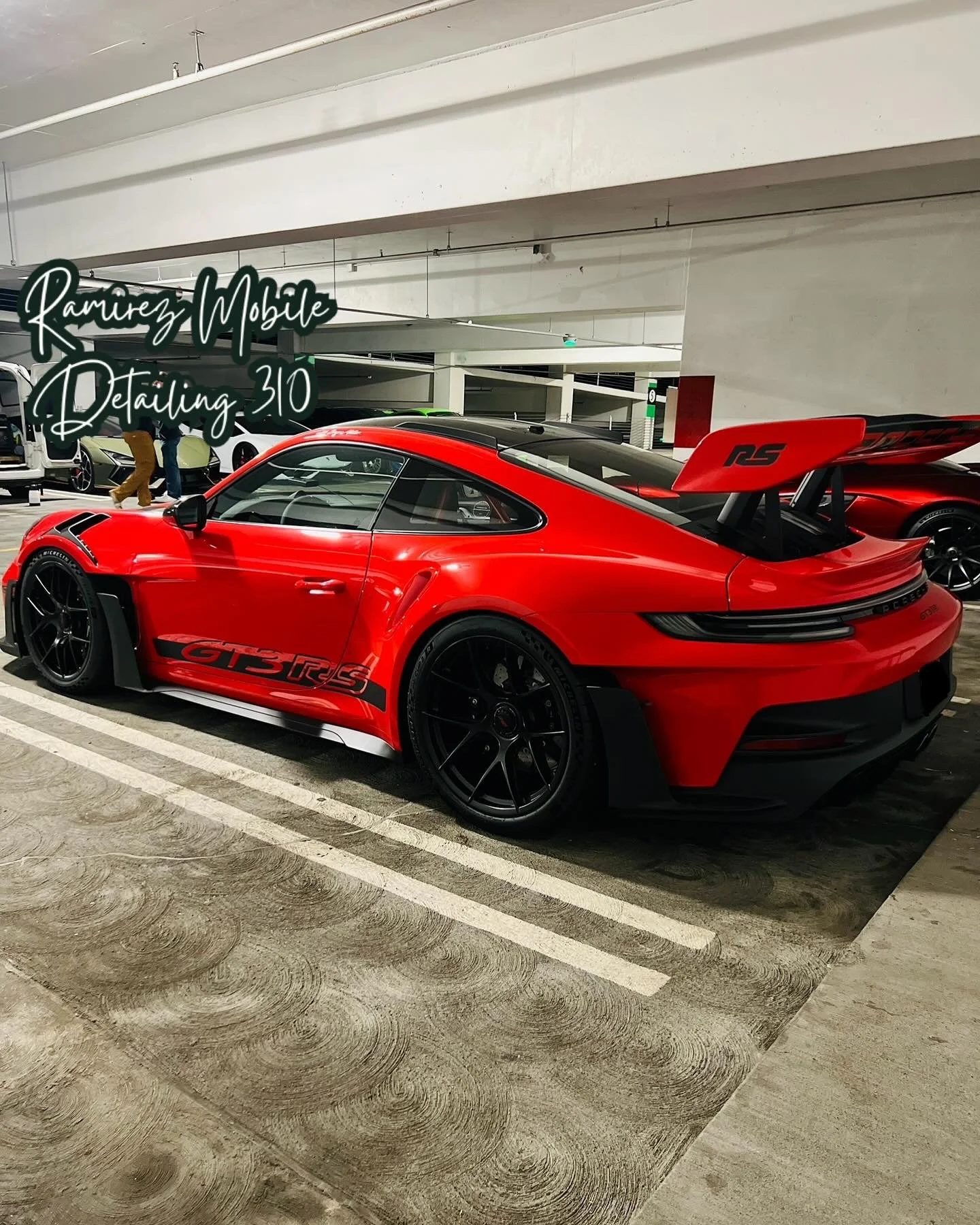 A red Porsche 911 GT3 RS parked in an indoor parking garage with a large rear wing and black wheels. Handwritten text in the top left corner says, "Ramirez Mobile Detailing 310."