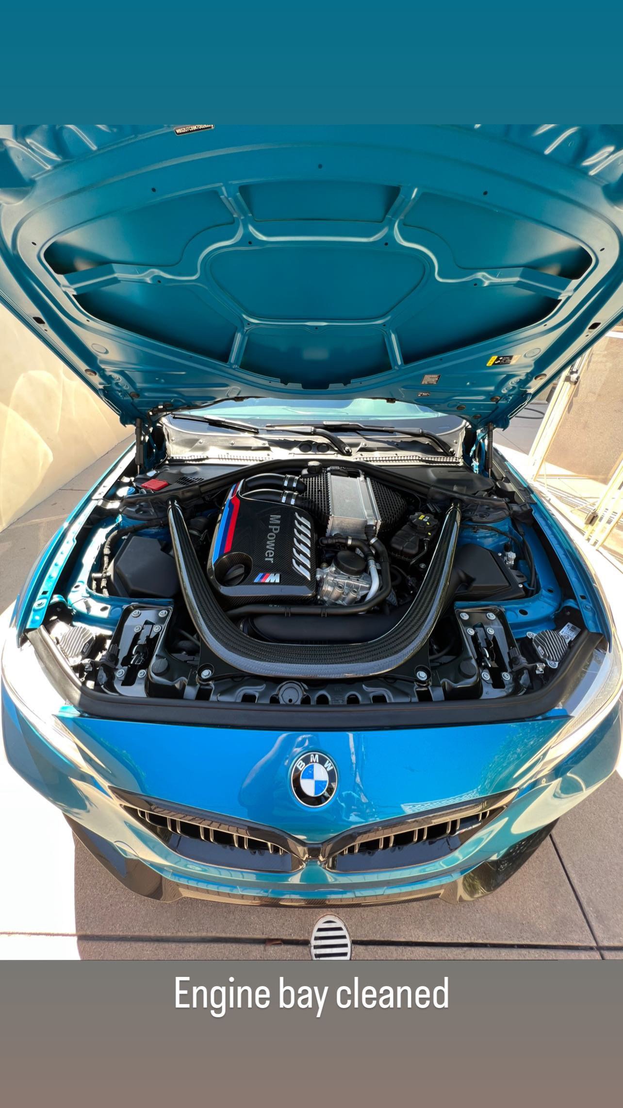 Blue BMW with open hood, showing a clean engine bay with an M Power engine and carbon fiber support brace, parked outside.