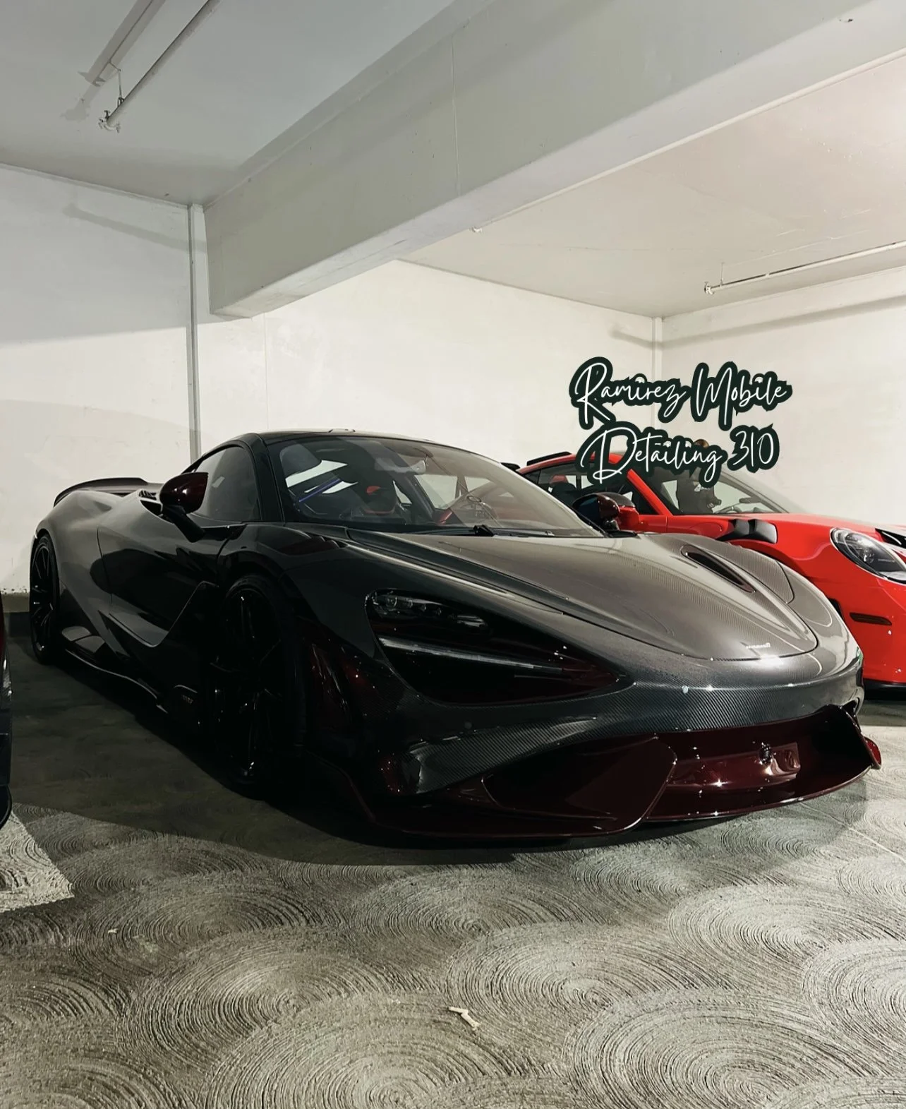 A black McLaren sports car with a carbon fiber hood parked indoors alongside a red sports car with a sign that reads 'Ramirez Mobile Detailing 310' in the background.
