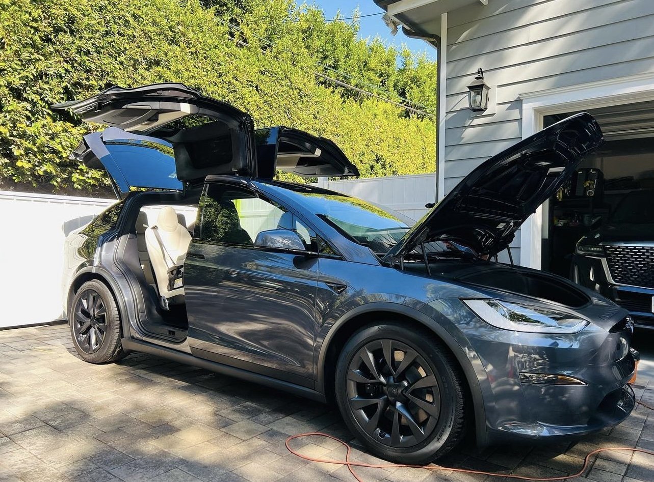 A dark gray Tesla Model Y electric vehicle with its front hood and rear liftgate open, parked in a driveway next to a white garage. The car's doors are open, and the interior with light-colored seats is visible. There is a black vehicle inside the garage and a cord on the ground.