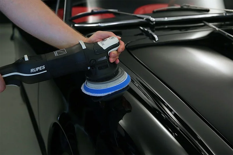 A person uses a Rupes electric polisher with a foam pad to polish the hood of a black car.