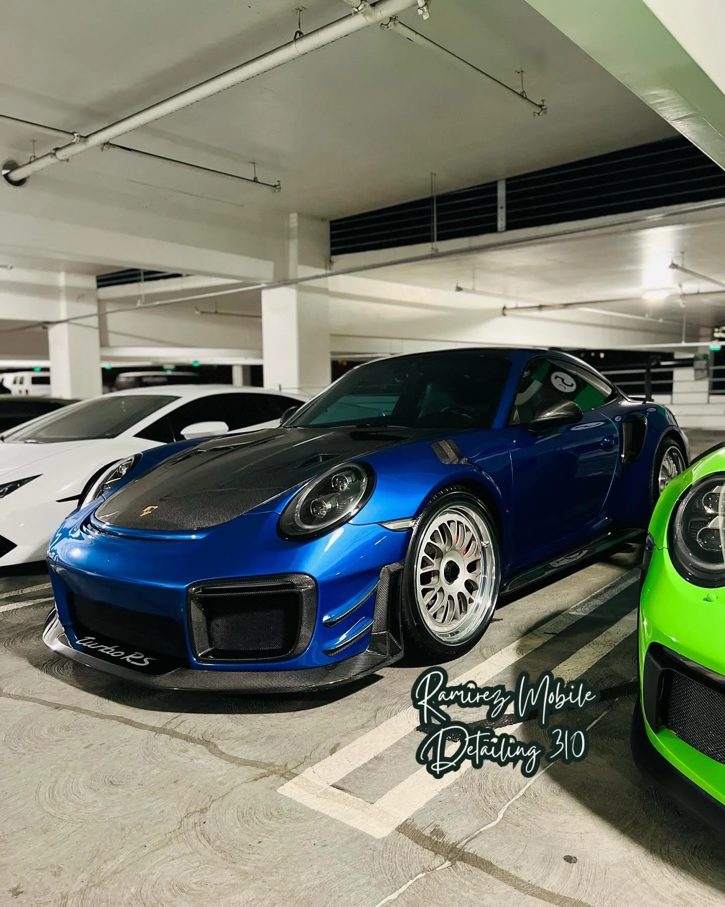A blue Porsche 911 Turbo 25 parked in an indoor parking garage, with a white car to its left and a green car to its right.