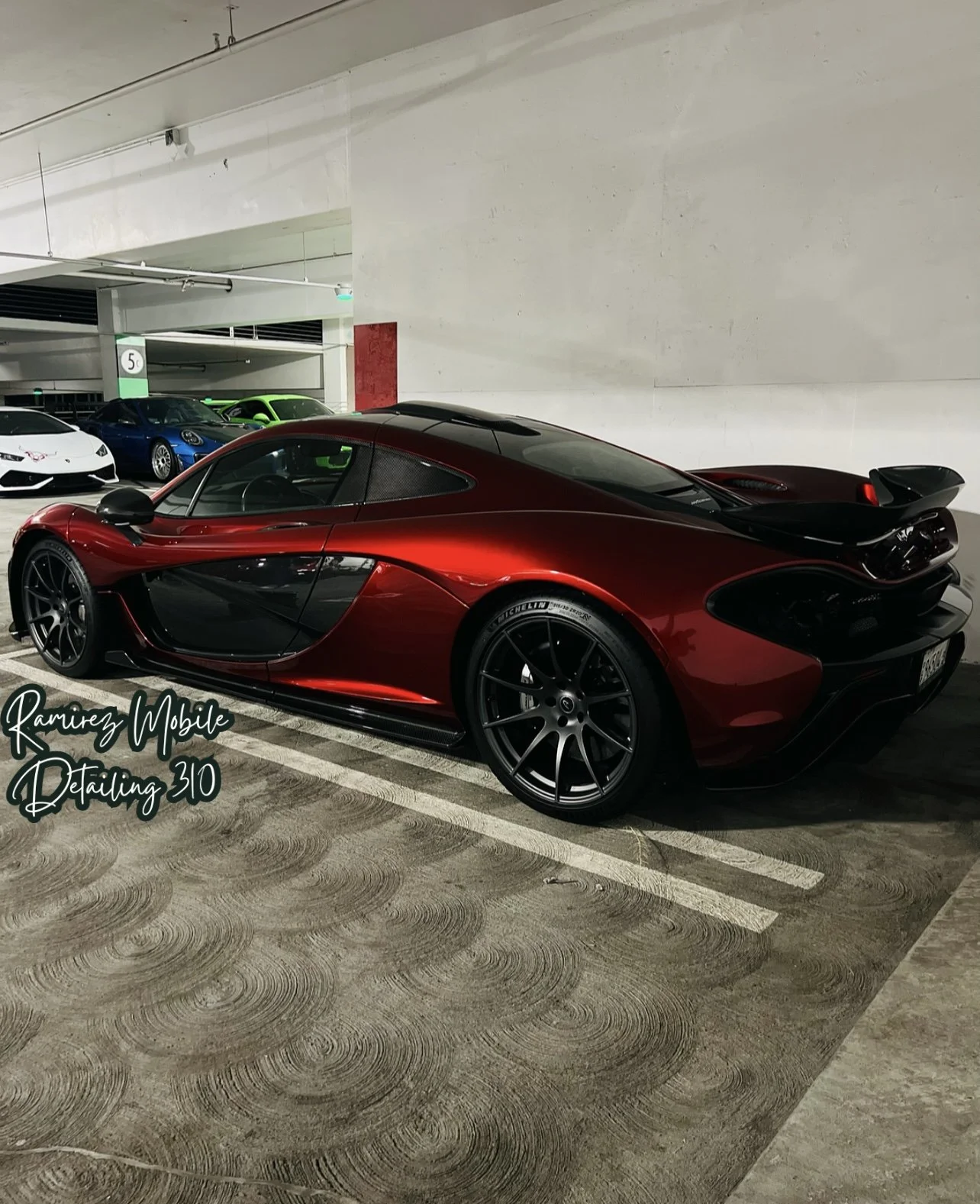 A red and black sports car parked in an indoor parking garage, with other cars visible in the background.