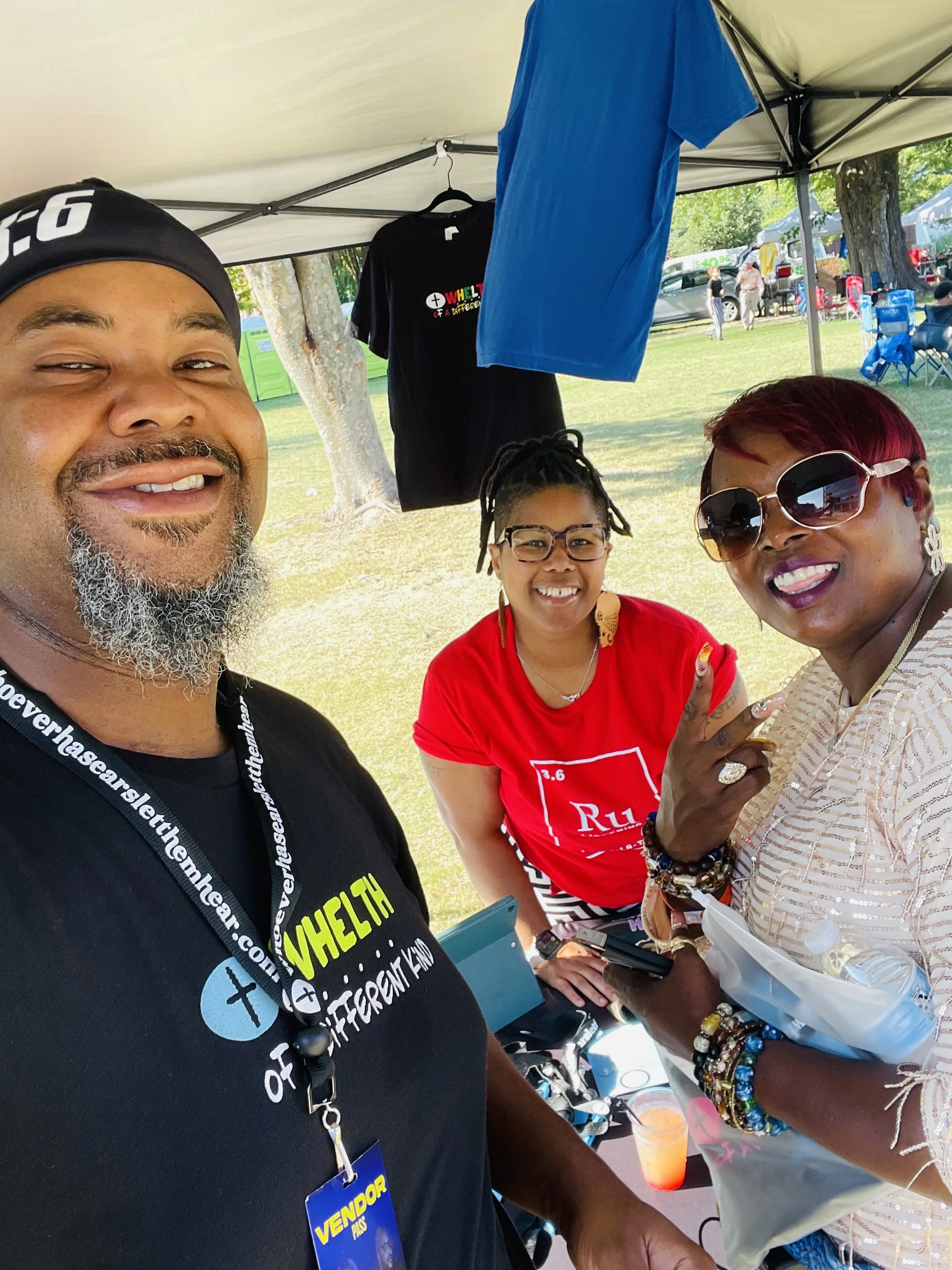 Three people smiling at an outdoor event under a canopy tent. The man on the left wears a black cap and a black t-shirt, the woman in the middle has short red hair and sunglasses, and the woman on the right has dreadlocks, glasses, and wears a white lace top. Clothes and other items hang from the tent, and there are tables, chairs, and other people in the background.