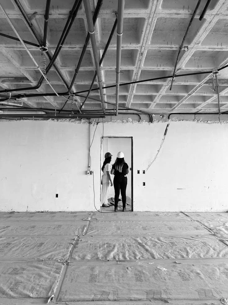 Two women wearing construction helmets in an unfinished room, looking at a doorway. The ceiling has exposed pipes and beams, and the floor is covered with construction materials.