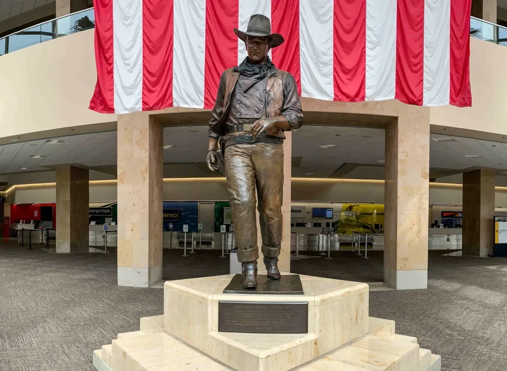 Bronze statue of a cowboy in a hat and boots, standing with one hand in his pocket, in an airport terminal with red, white, and blue flag decorations behind it.