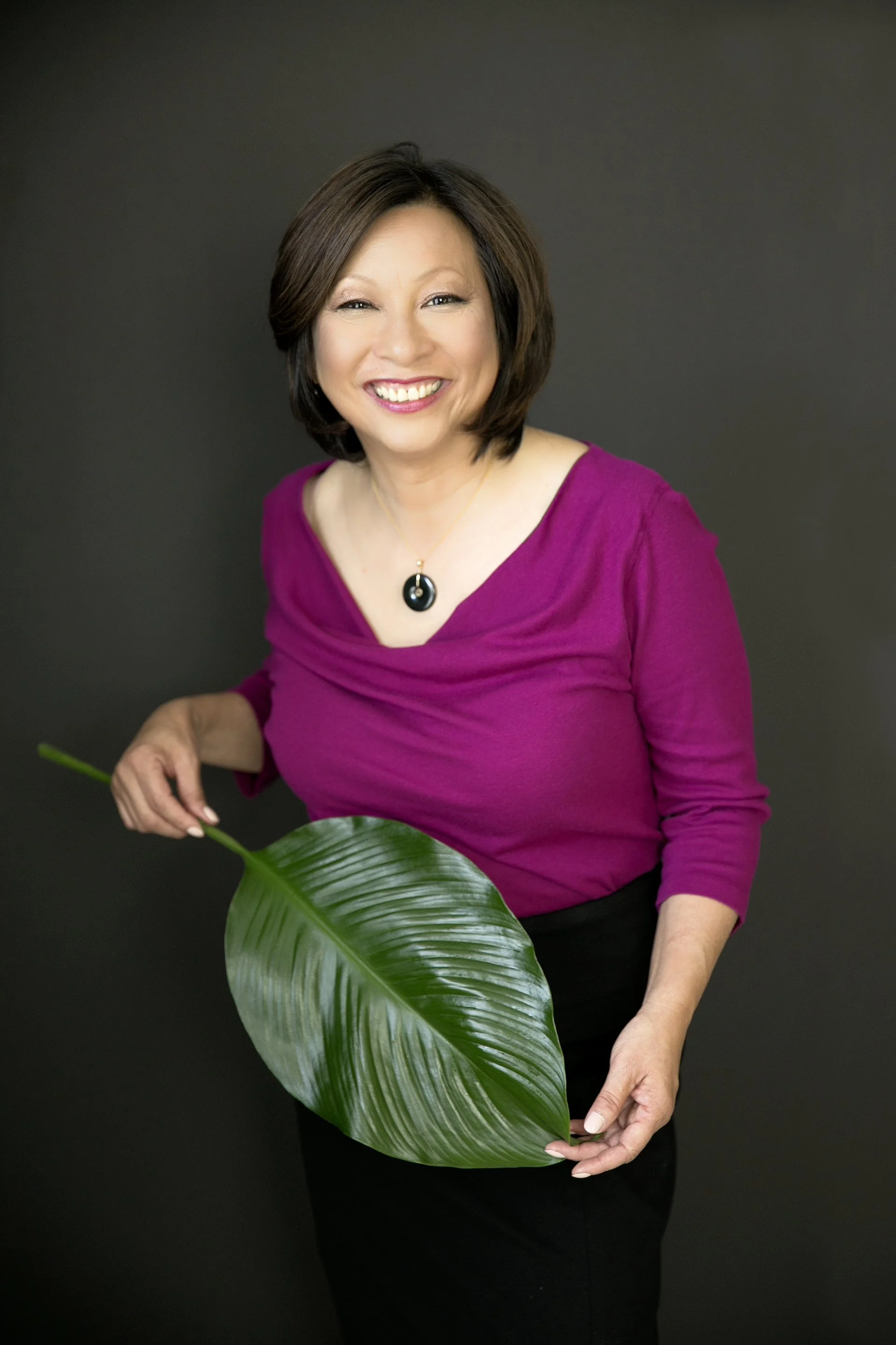 Connie Hom in a magenta top, holding a large glossy tropical leaf against a dark gray backdrop.