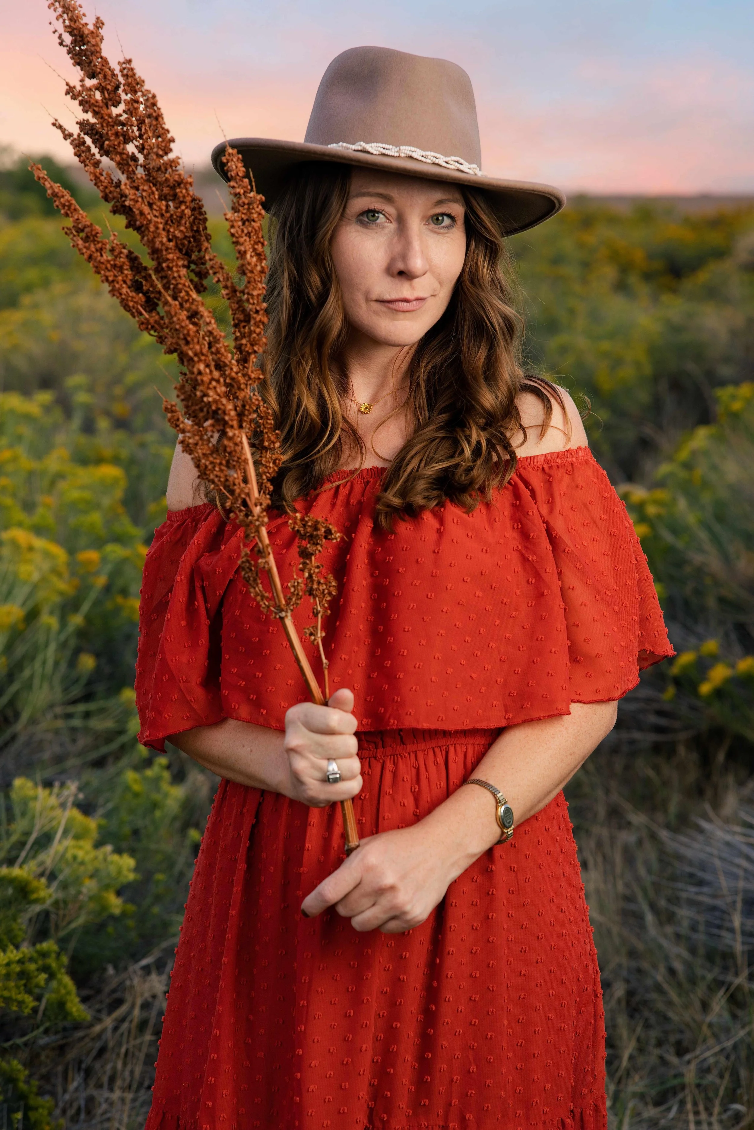 Smiling woman in a rust dress holding white wildflowers in front of yellow rabbitbrush at sunset