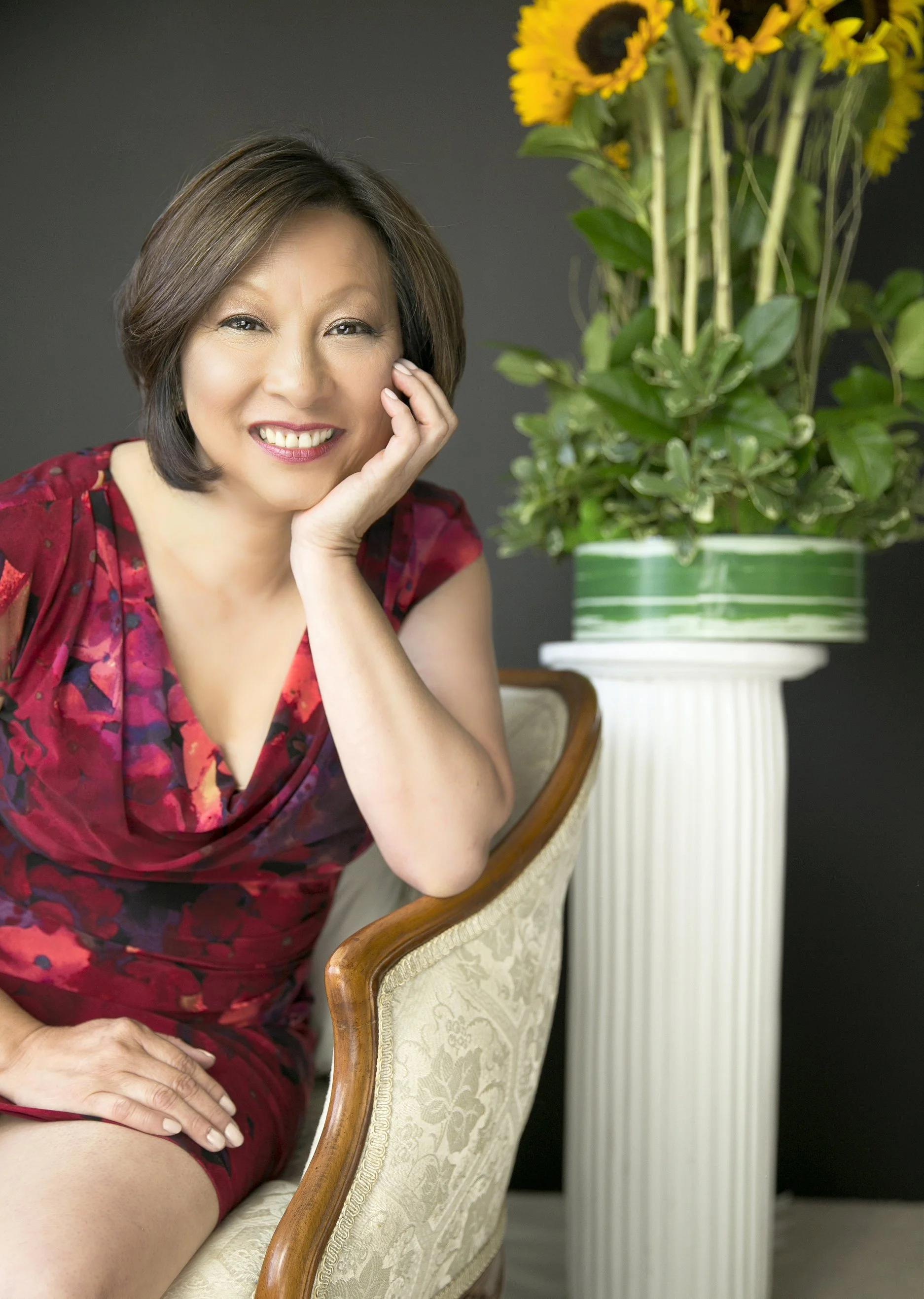 Connie Hom in a red floral dress, seated beside a tall sunflower arrangement during her personal branding session in Richmond, VA.