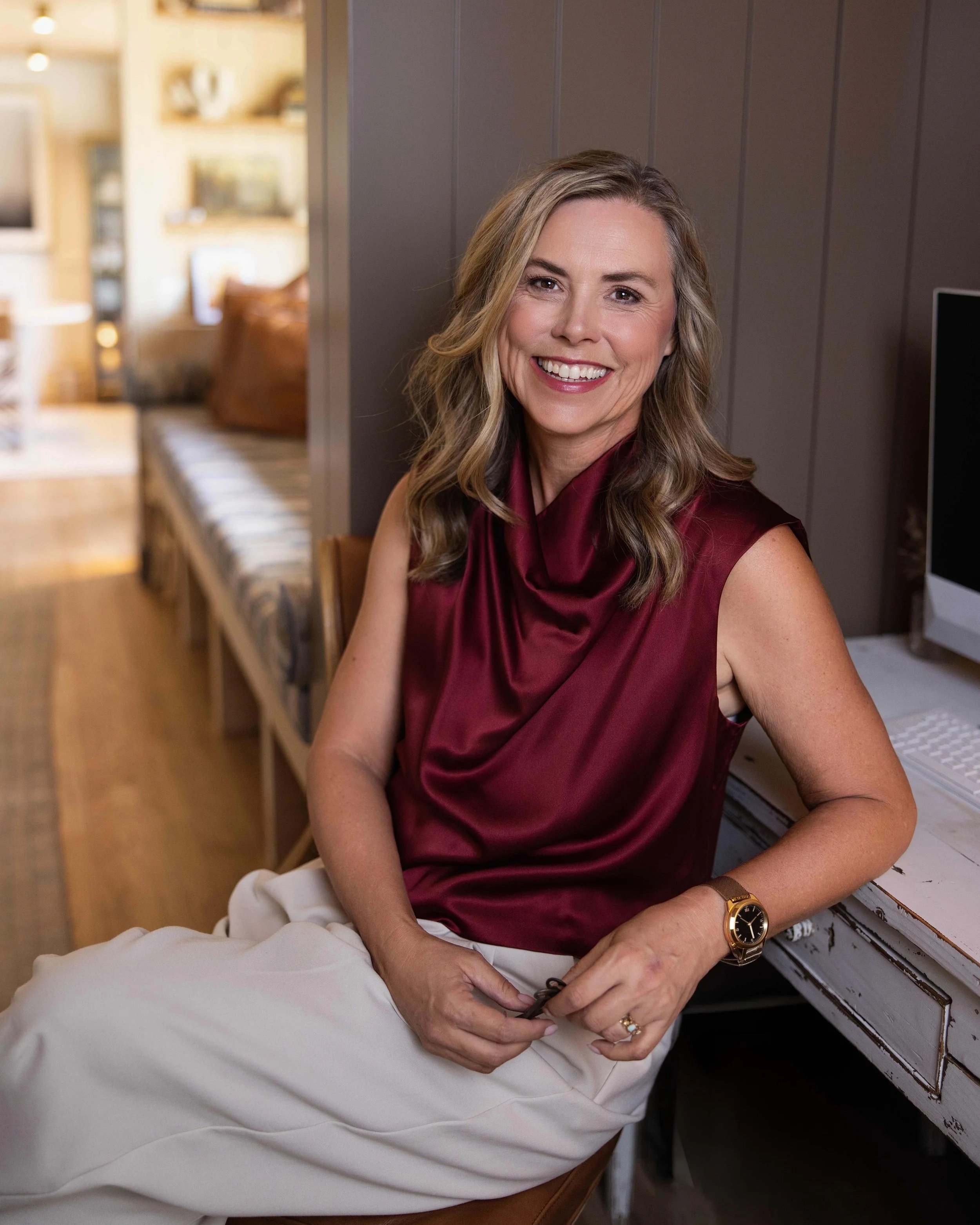 Kim Brundage sitting at a computer desk wearing a burgundy silk top and cream pants.