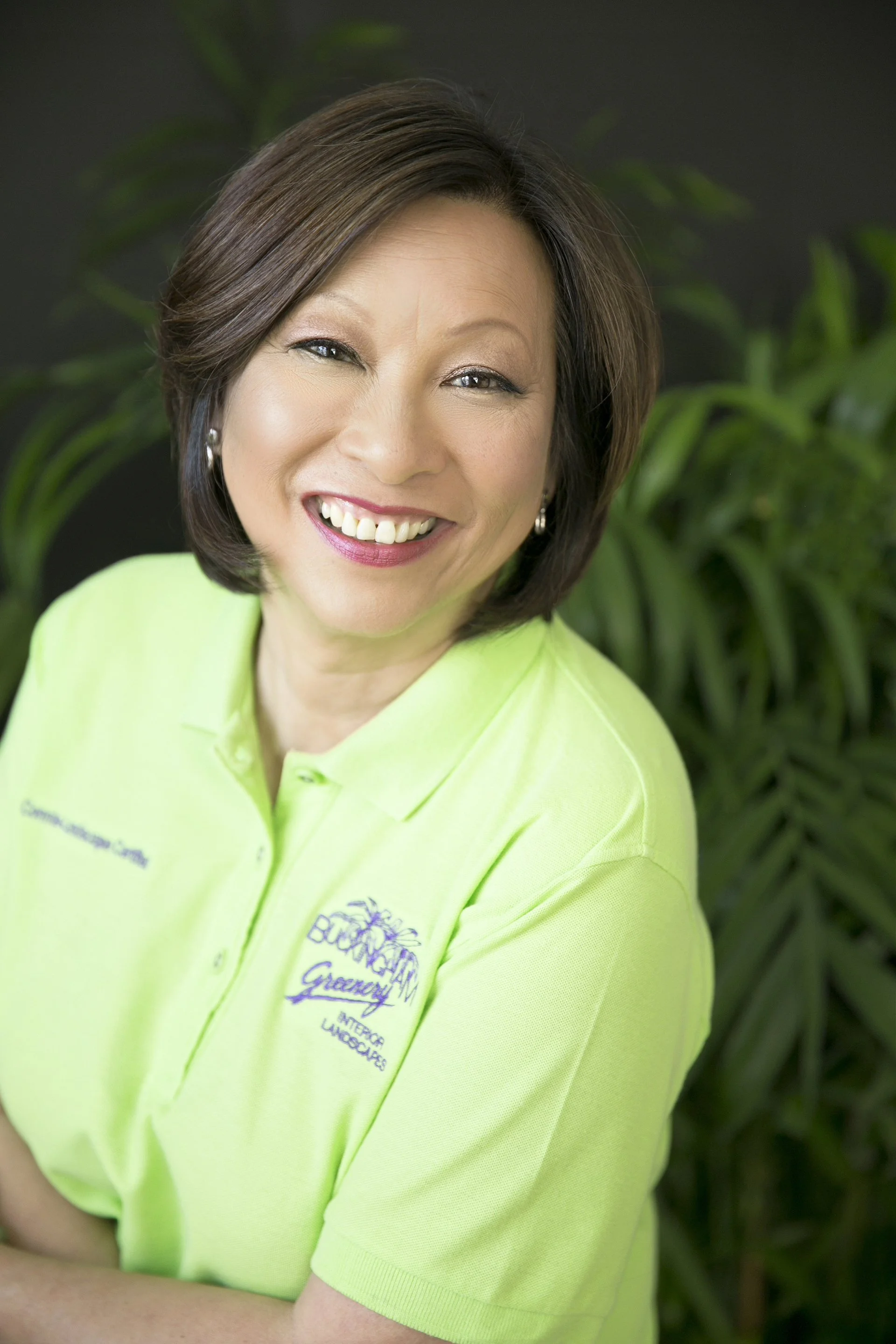 Connie Hom smiling in her lime green Buckingham Greenery polo, photographed in front of tropical foliage.