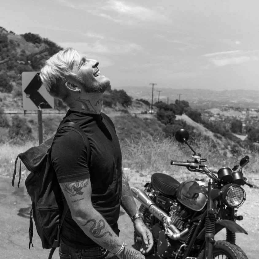 A smiling man with tattoos on his arms and neck, standing next to a motorcycle on a road in a hilly area with a scenic view in Portland, Oregon.