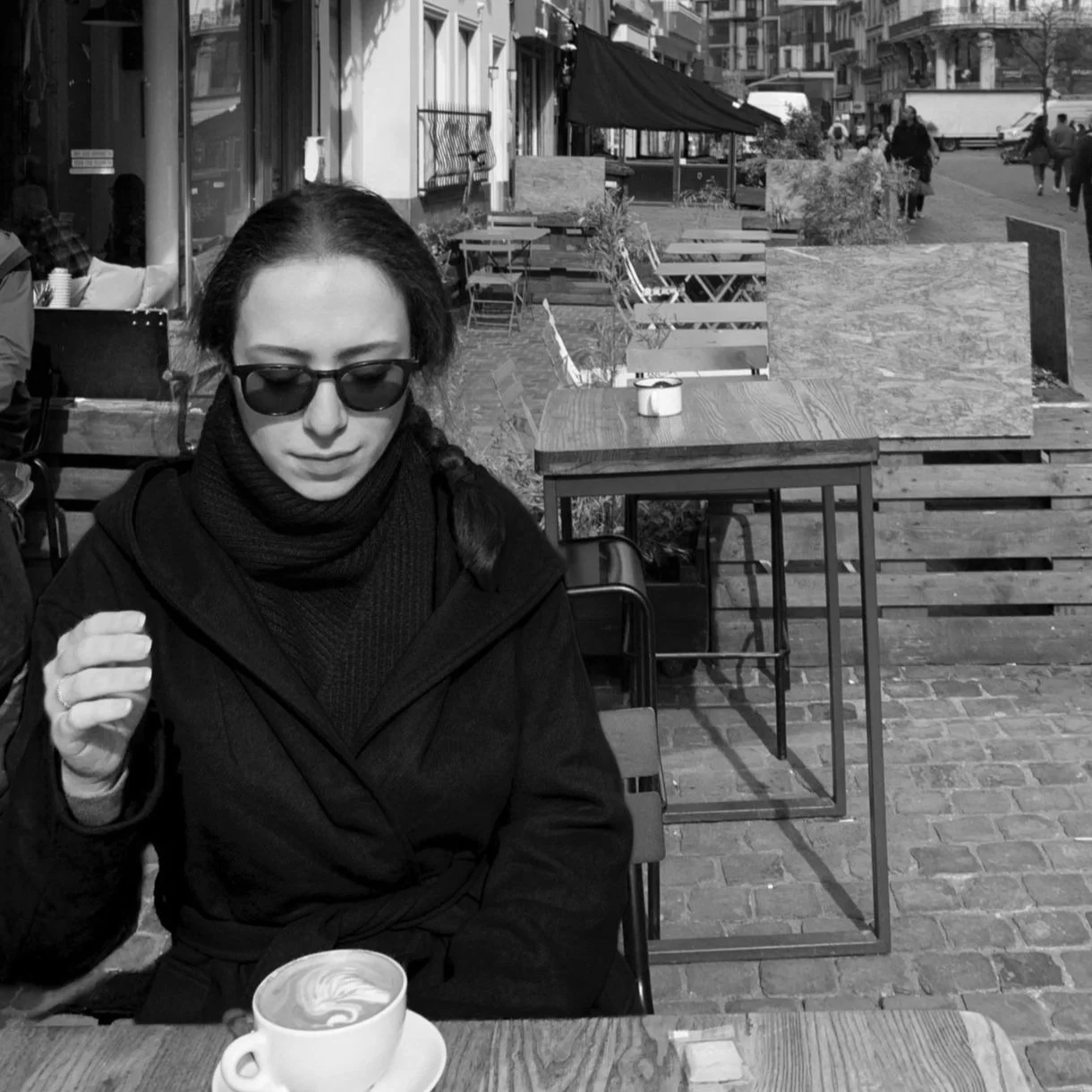 A woman sitting outdoors at a café with a cappuccino in front of her. There are empty chairs and tables behind her, and some people walking along the street in the background in Portland, Oregon.