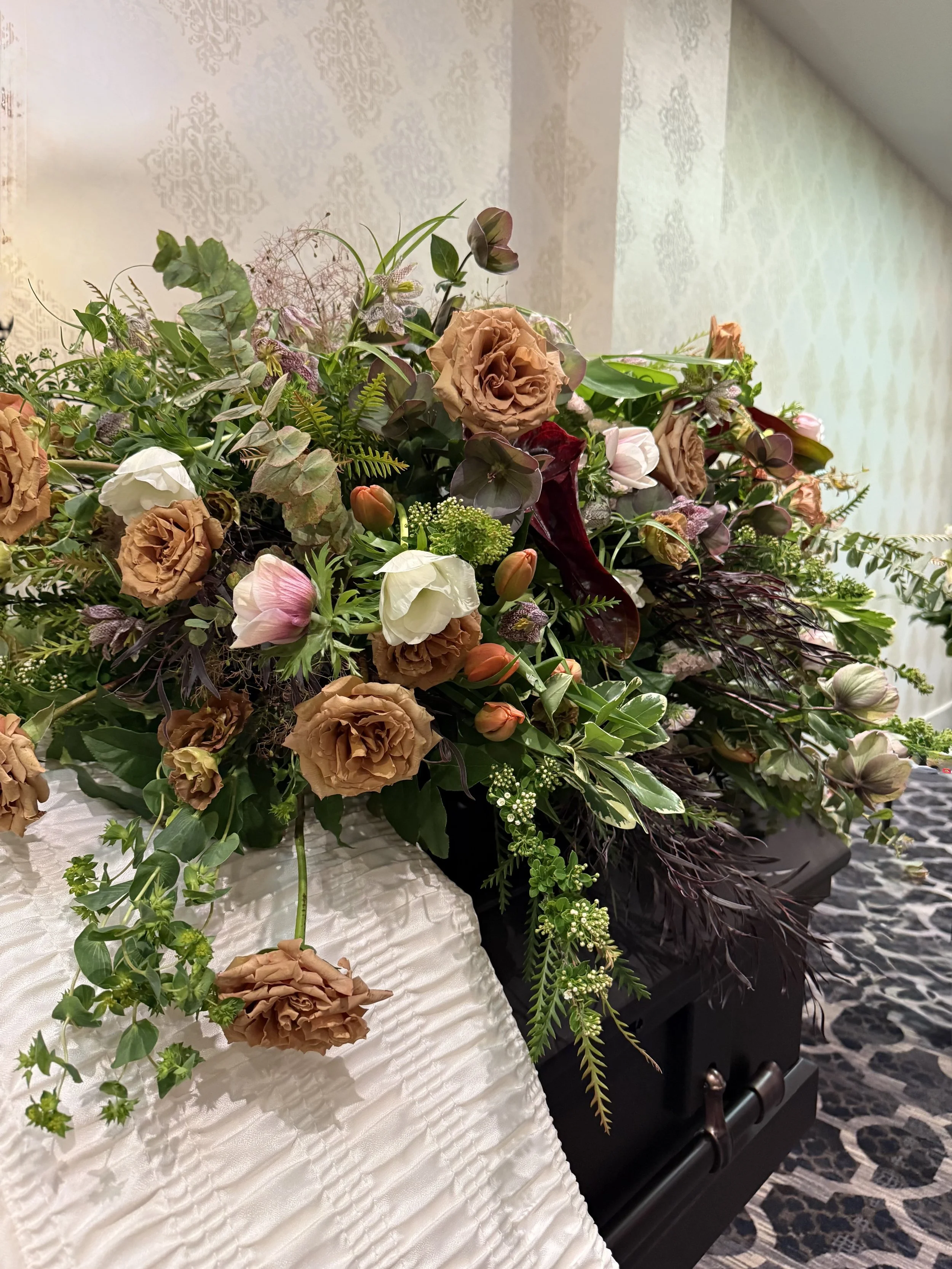 A large flower arrangement with cream, white, and dark red flowers, green leaves, and lilac accents displayed on a table with a white cloth, and a patterned wall in the background.