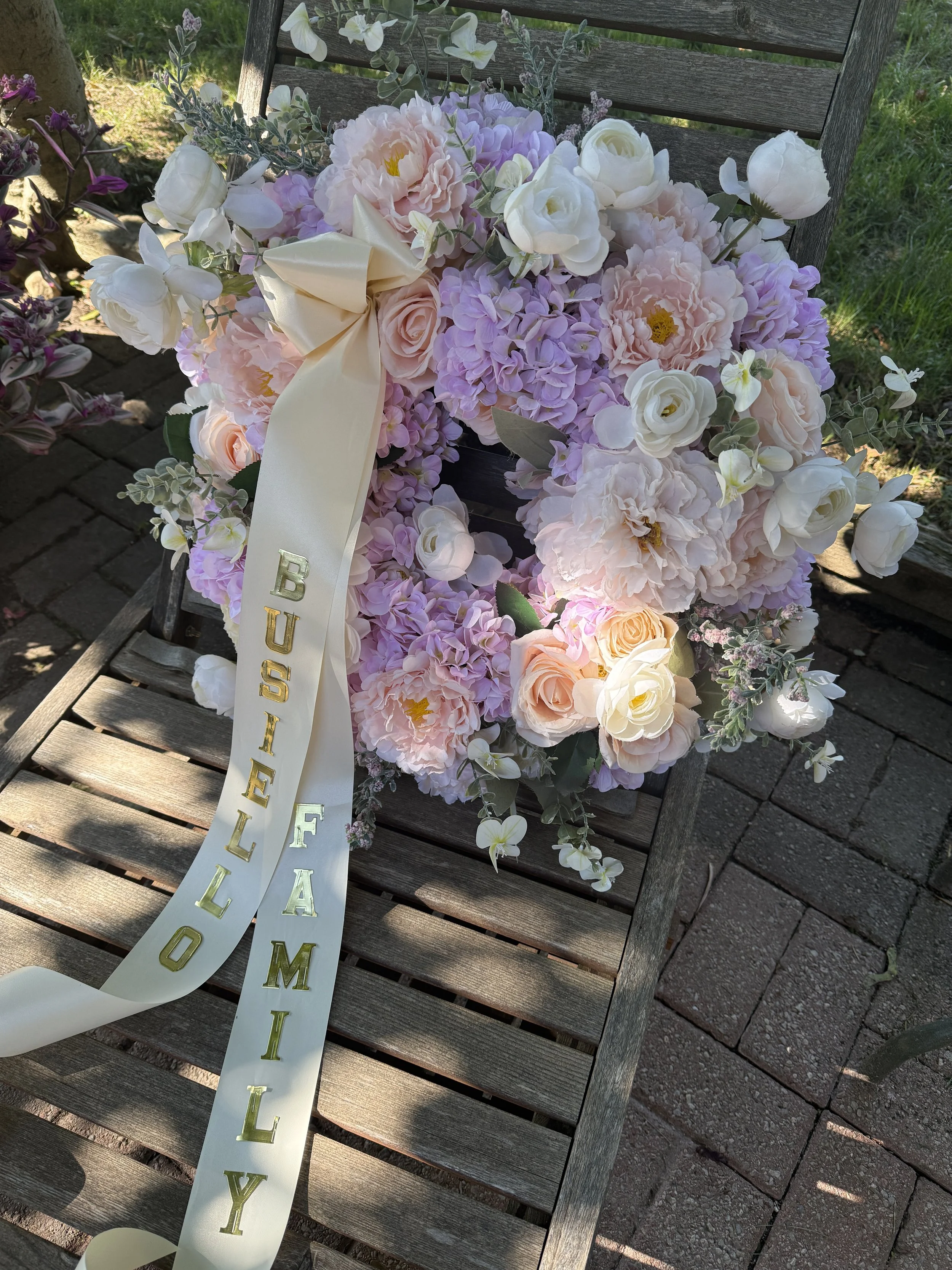 A floral arrangement with pink and white roses, purple hydrangeas, white orchids, and greenery on a wooden bench outdoors.