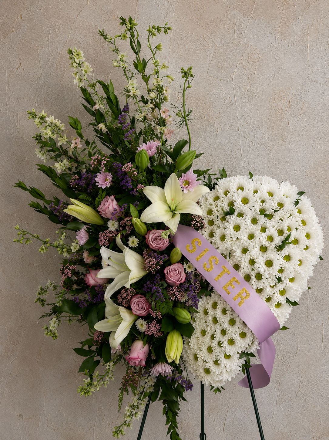 A floral funeral arrangement with white lilies, pink roses, purple flowers, and white daisies, decorated with a lavender ribbon that says 'SISTER.'