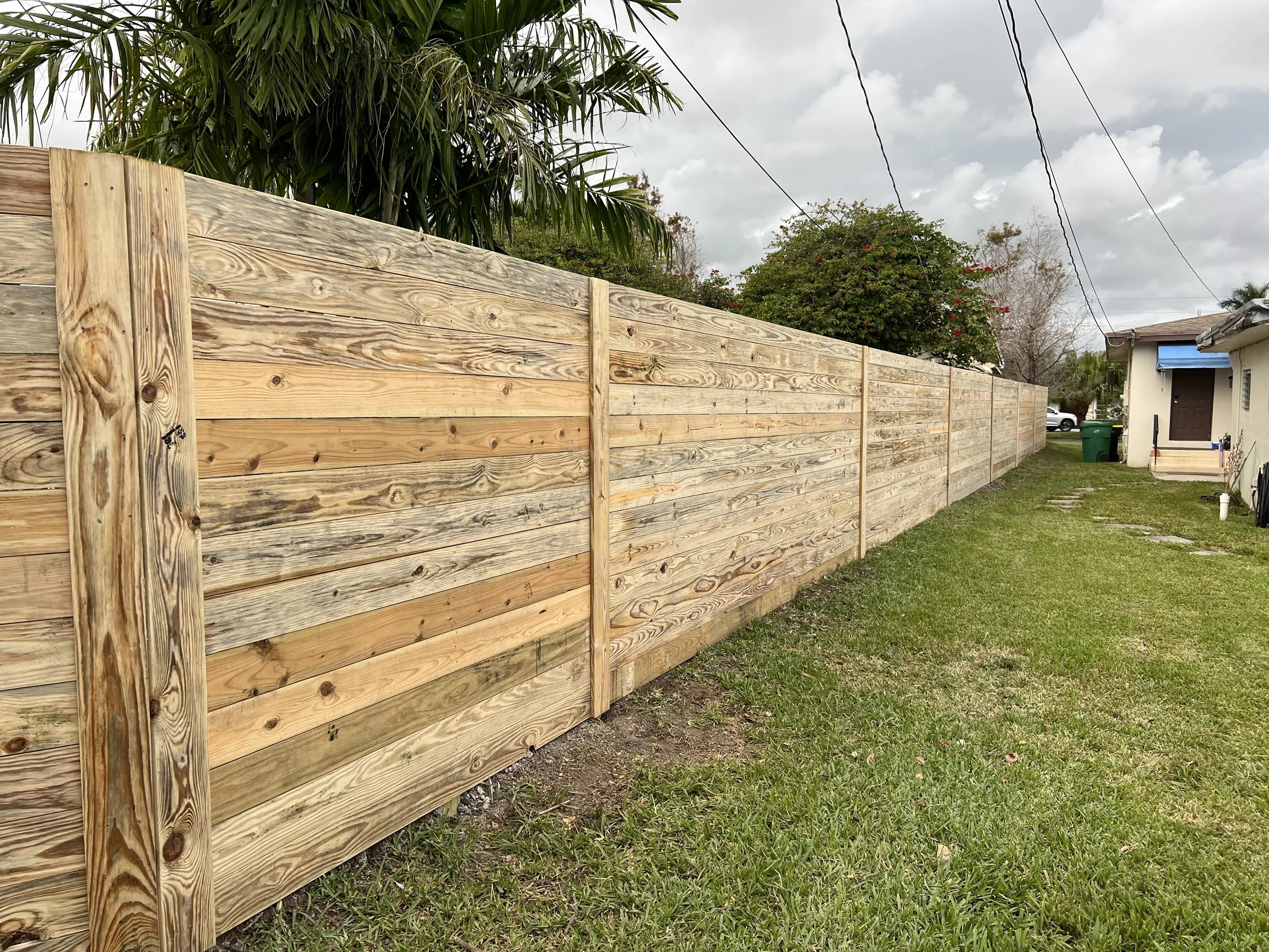 A newly installed wooden privacy fence runs along a grassy backyard, with houses and trees in the background under a partly cloudy sky.