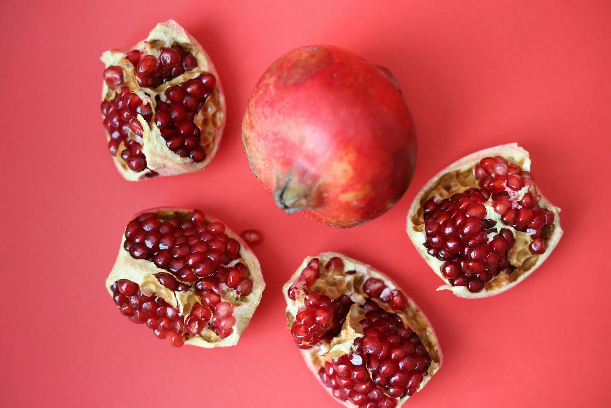 Whole pomegranate fruit surrounded by five open pomegranate halves on a pink background.