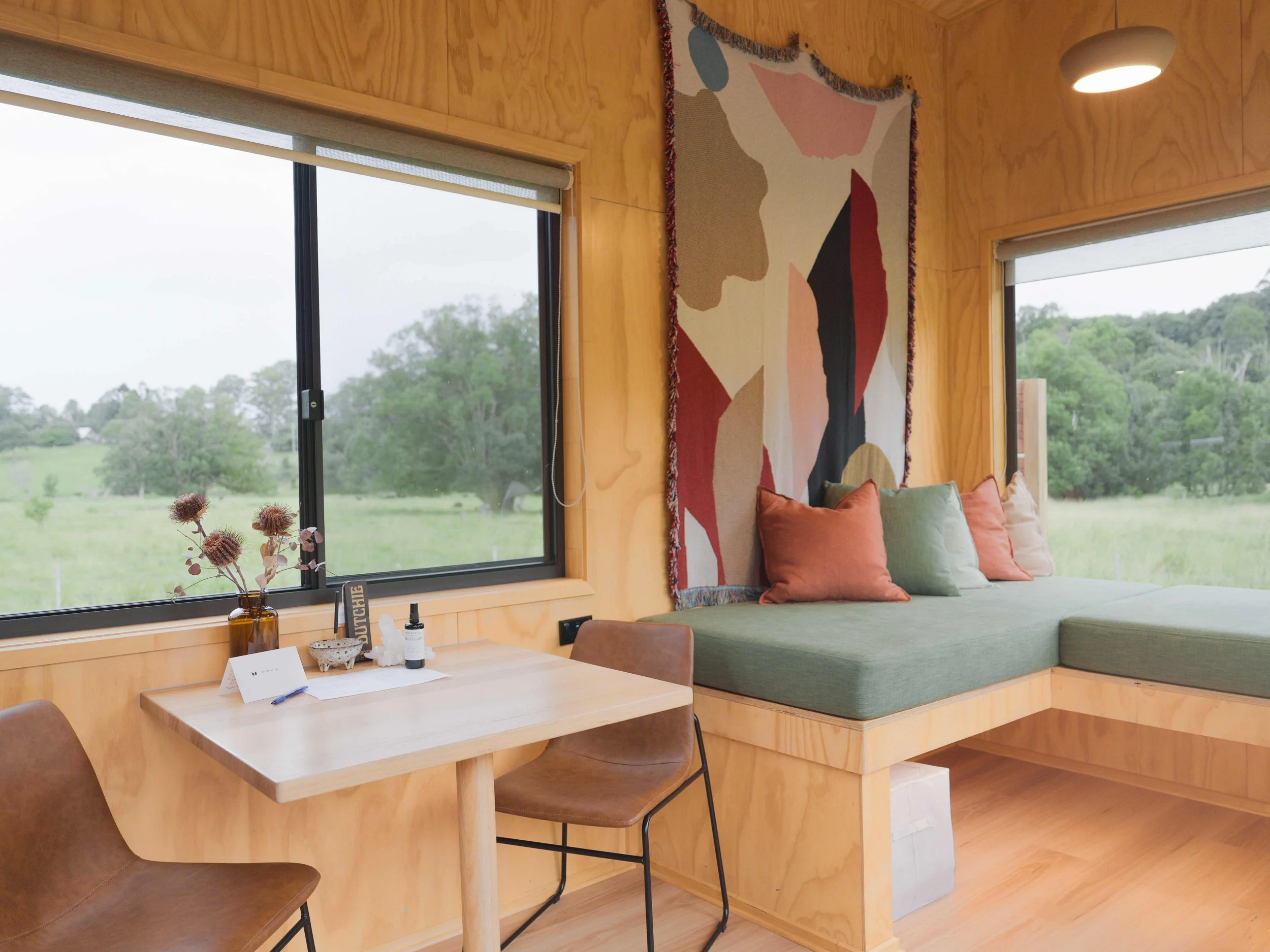 Cozy corner of a room featuring a large window, a wooden bench with green cushions and multiple pillows, an abstract wall hanging, and a small table with dried flowers, bottles, and papers, overlooking a lush green landscape.