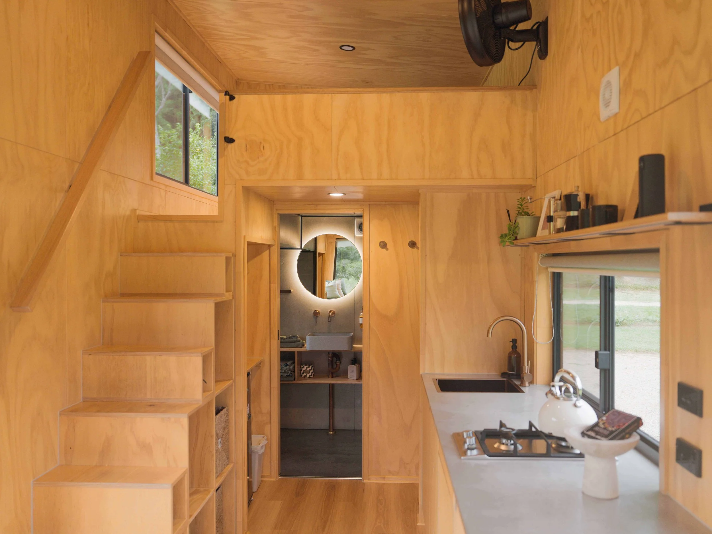 Interior view of a small wooden kitchen with a staircase on the left, a window above the sink, a countertop on the right with a kettle and a bowl, a small black fan, and a bathroom with a round mirror visible through an open doorway at the back.