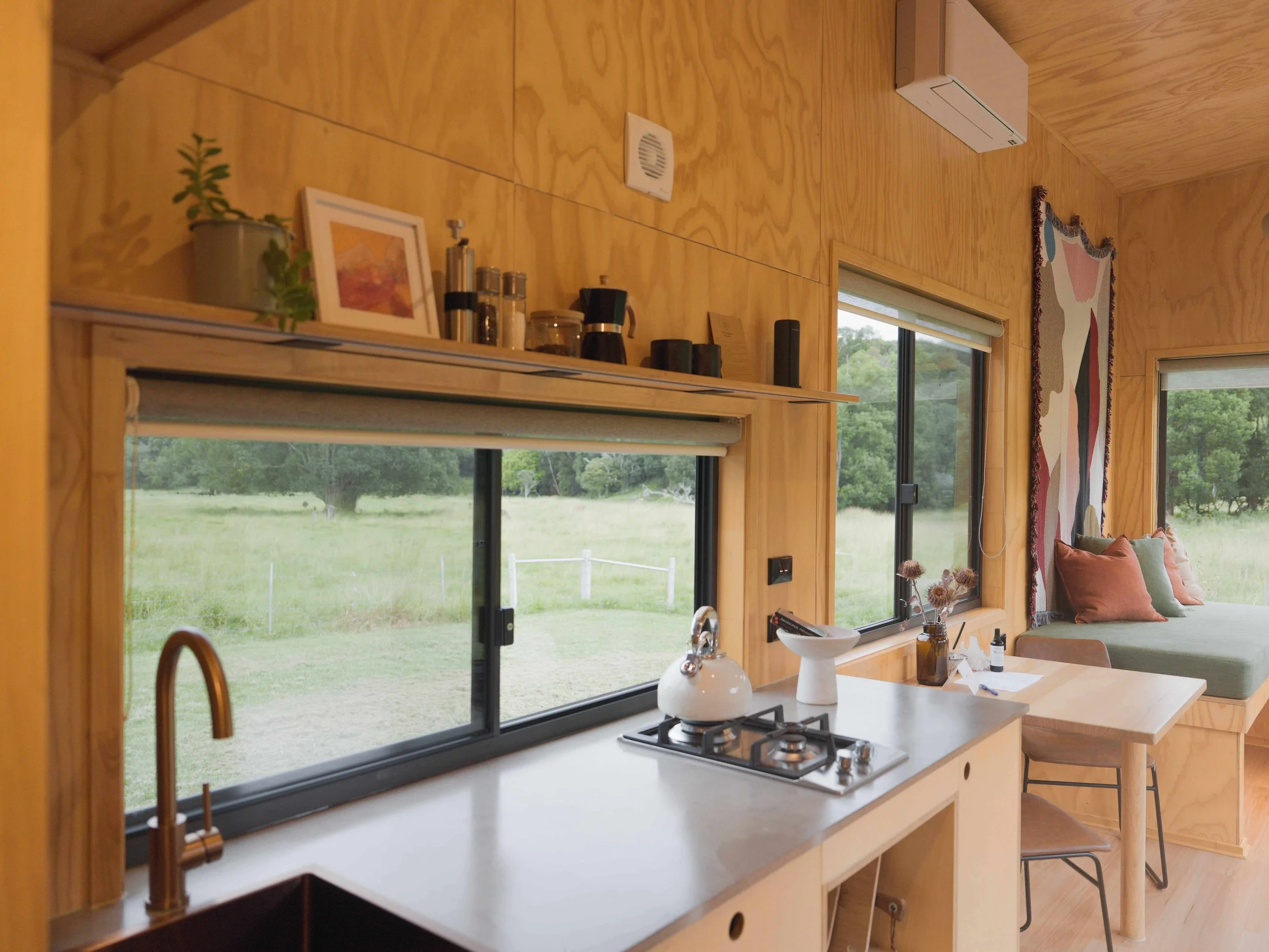 Interior of a cozy wood-paneled kitchen with a view of a green outdoor landscape through windows. The kitchen features a white countertop with a stove, a copper faucet, and a small sink. There are decorative items on the shelf above, including a smal