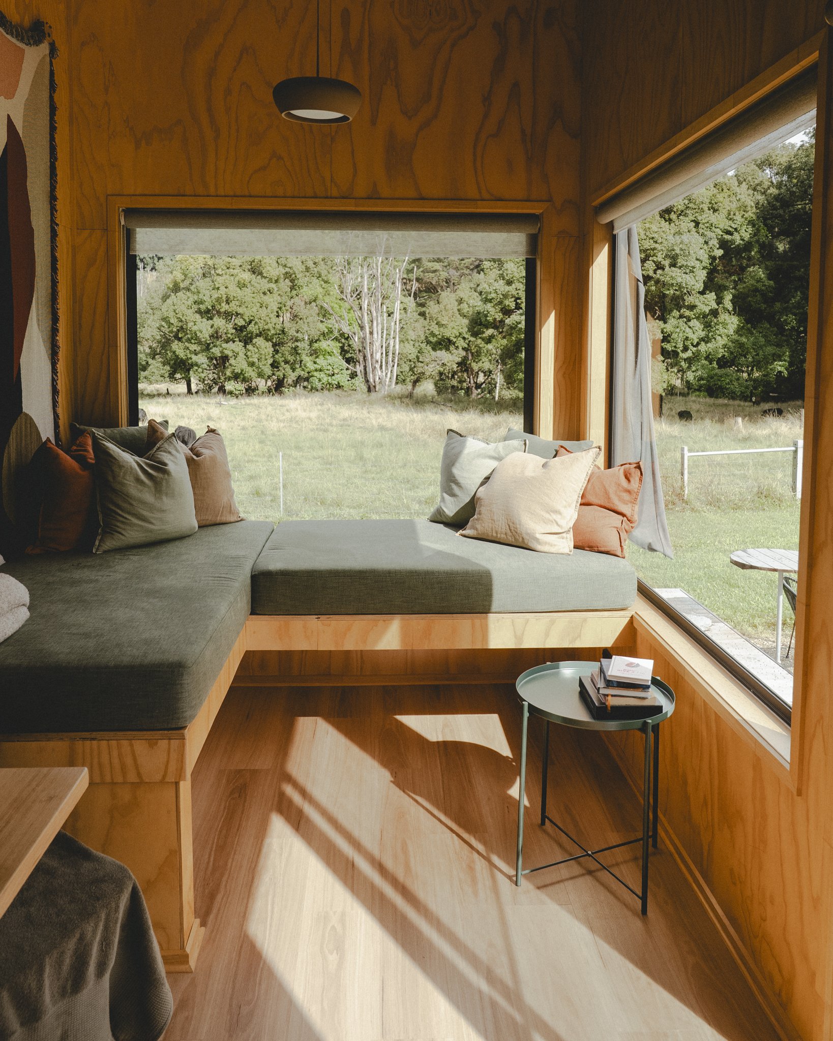 Cozy corner inside a wooden cabin with large window seating, cushions, and small side table with books, overlooking a green grassy landscape with trees.