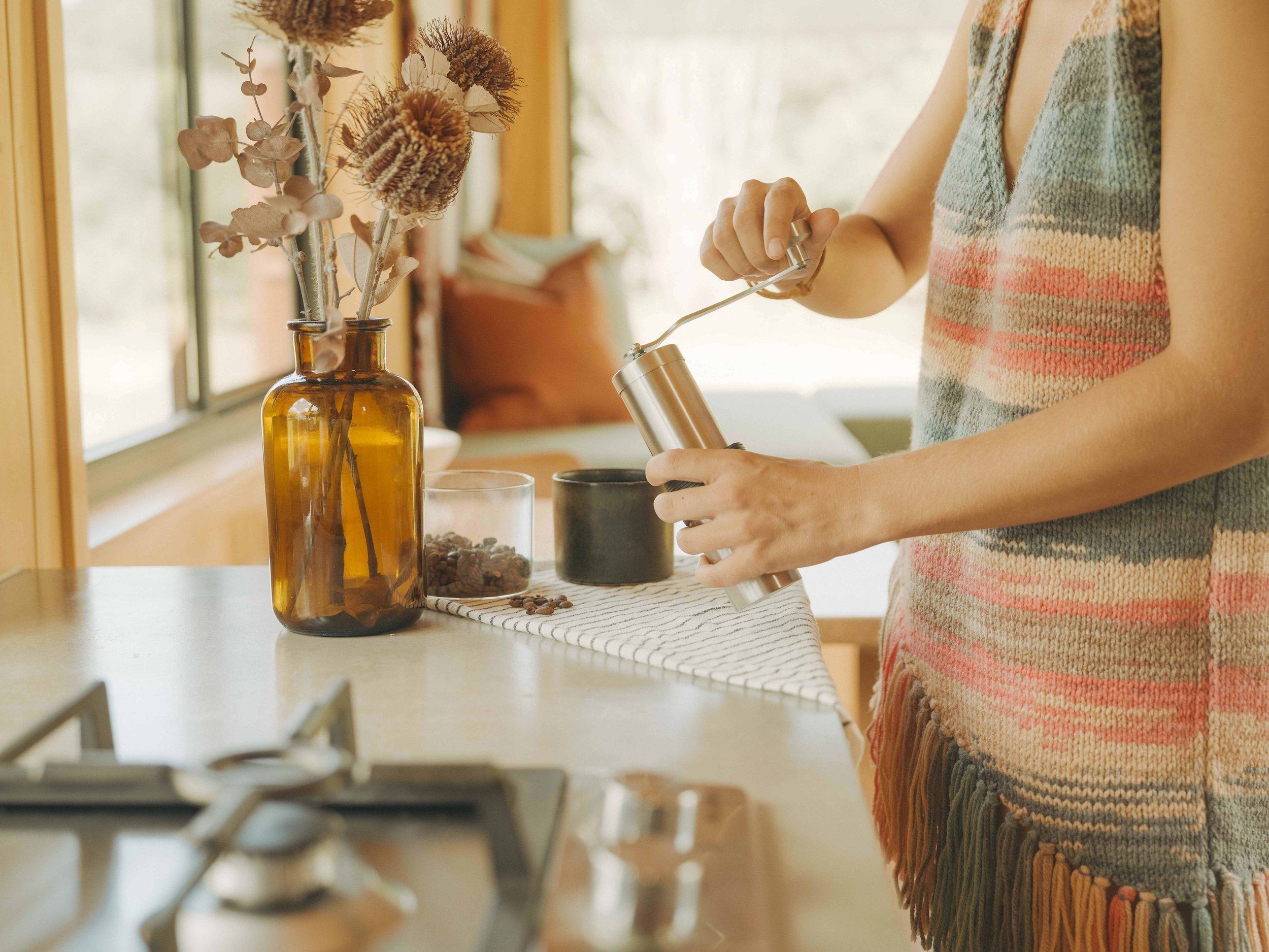 Person using a manual coffee grinder in a bright kitchen with a window, a vase with dried flowers, a glass container with coffee beans, and a black mug on the counter.
