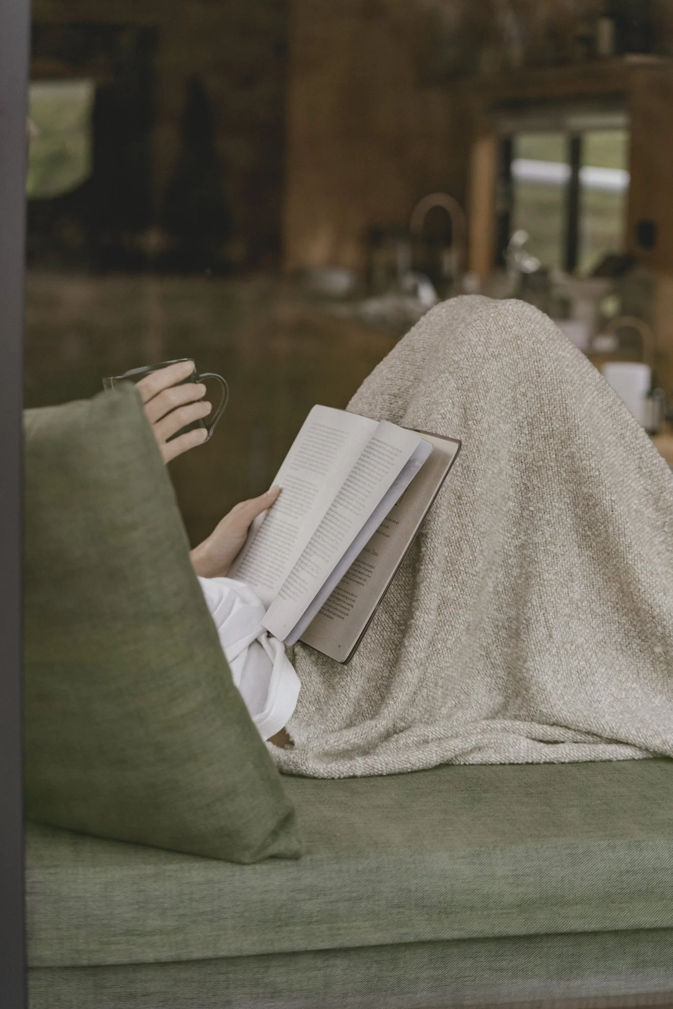 Person sitting on a couch with legs up, reading a book and holding a mug, in a cozy tiny home in Byron Bay Hinterland.