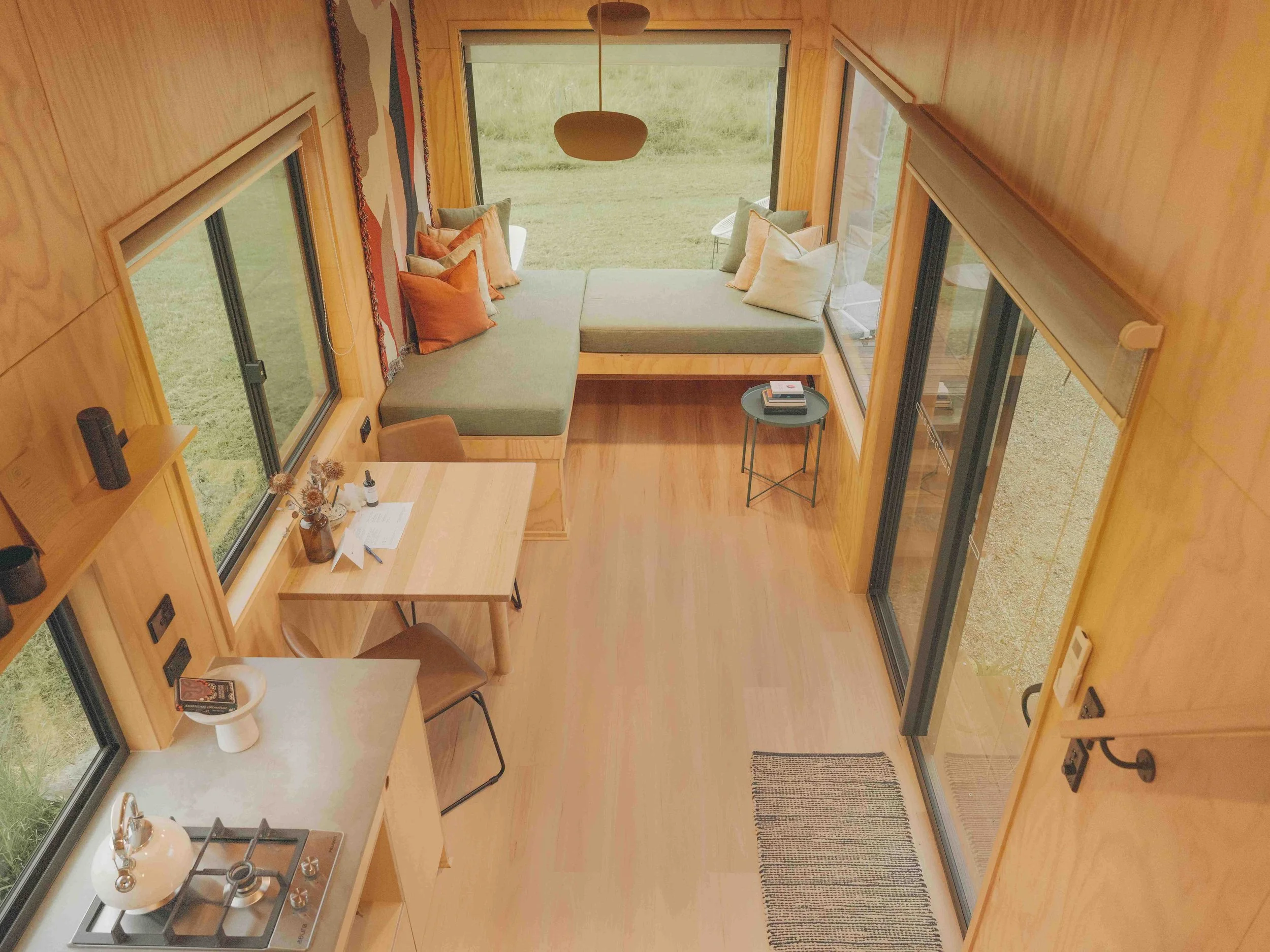 A cozy, modern tiny house interior with wooden walls and large windows. It features a fold-out sofa with multiple cushions near the wall, a small side table with books, a dining area with a wooden table and chairs, and a kitchenette with a stove and 