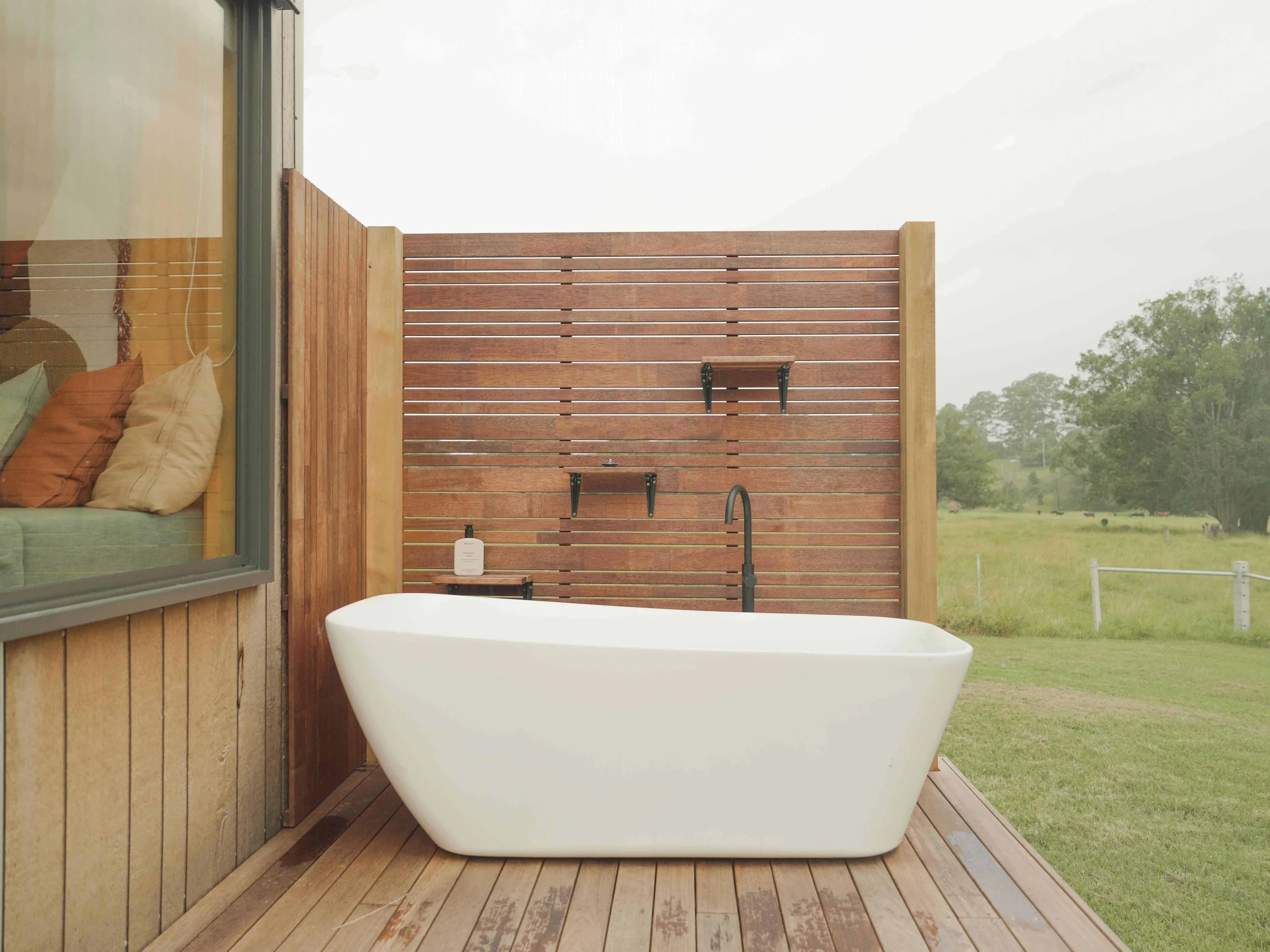 Outdoor bathtub on wooden deck with a wood privacy wall and view of grassy field and trees.