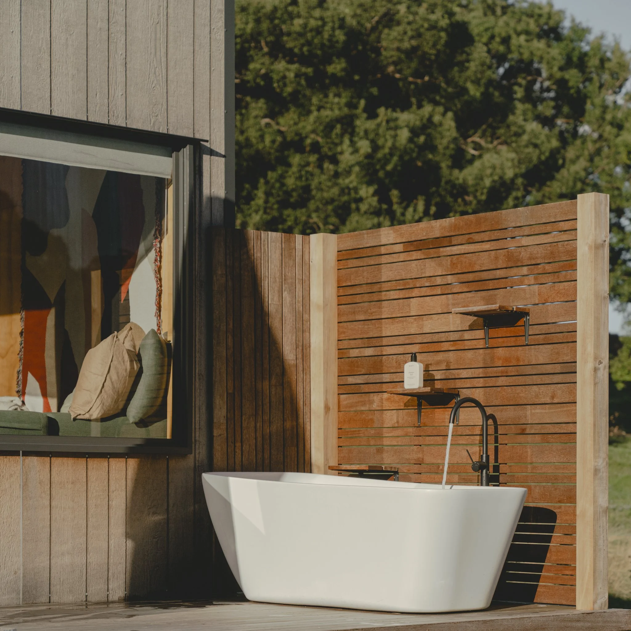 Outdoor bathtub next to a wooden privacy fence with mounted shelves, set against a background of greenery and a house window with visible pillows and decor inside.