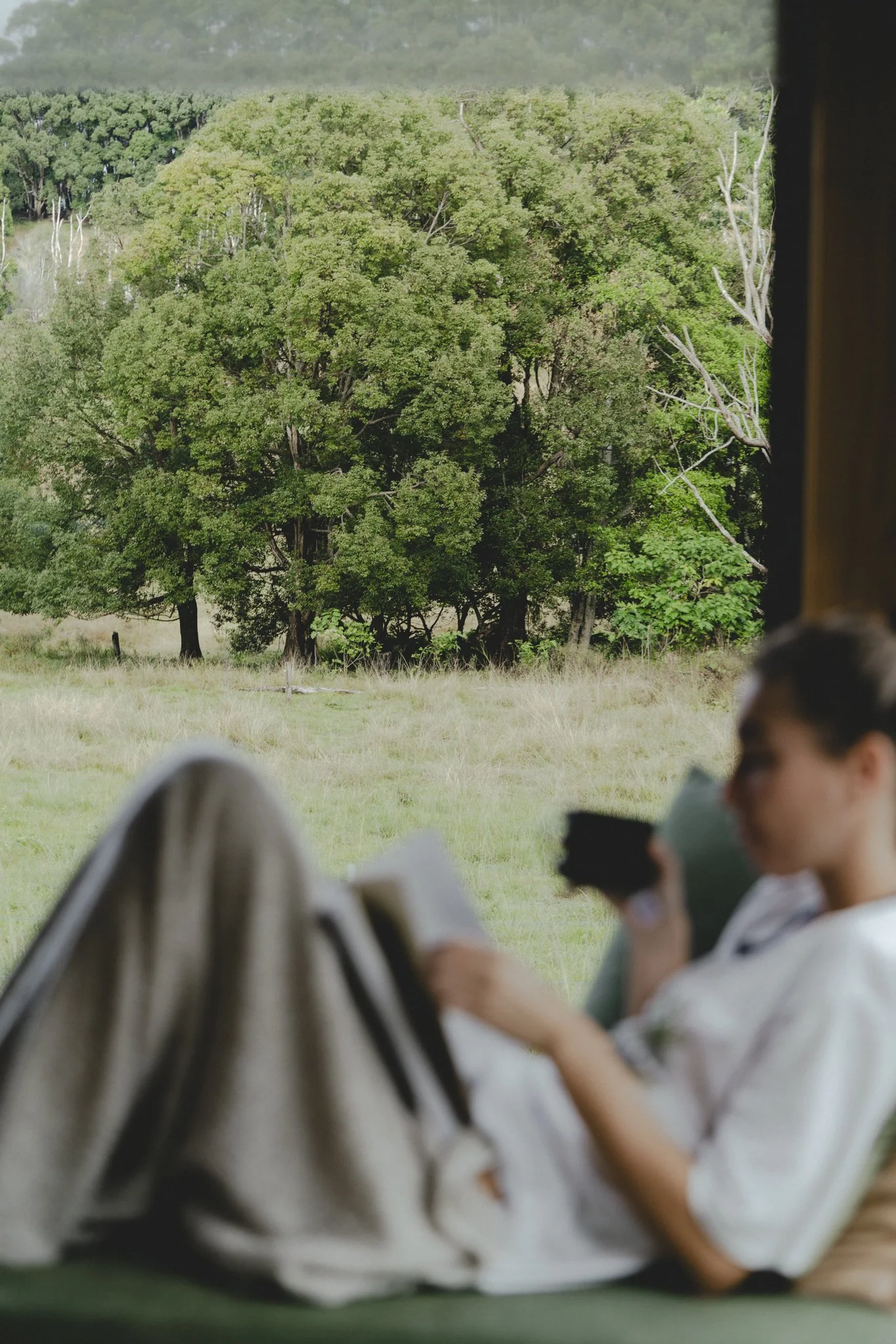 A woman sitting inside a room, reading a book and drinking coffee, with a view of green trees and grass through a window of an off-grid tiny home