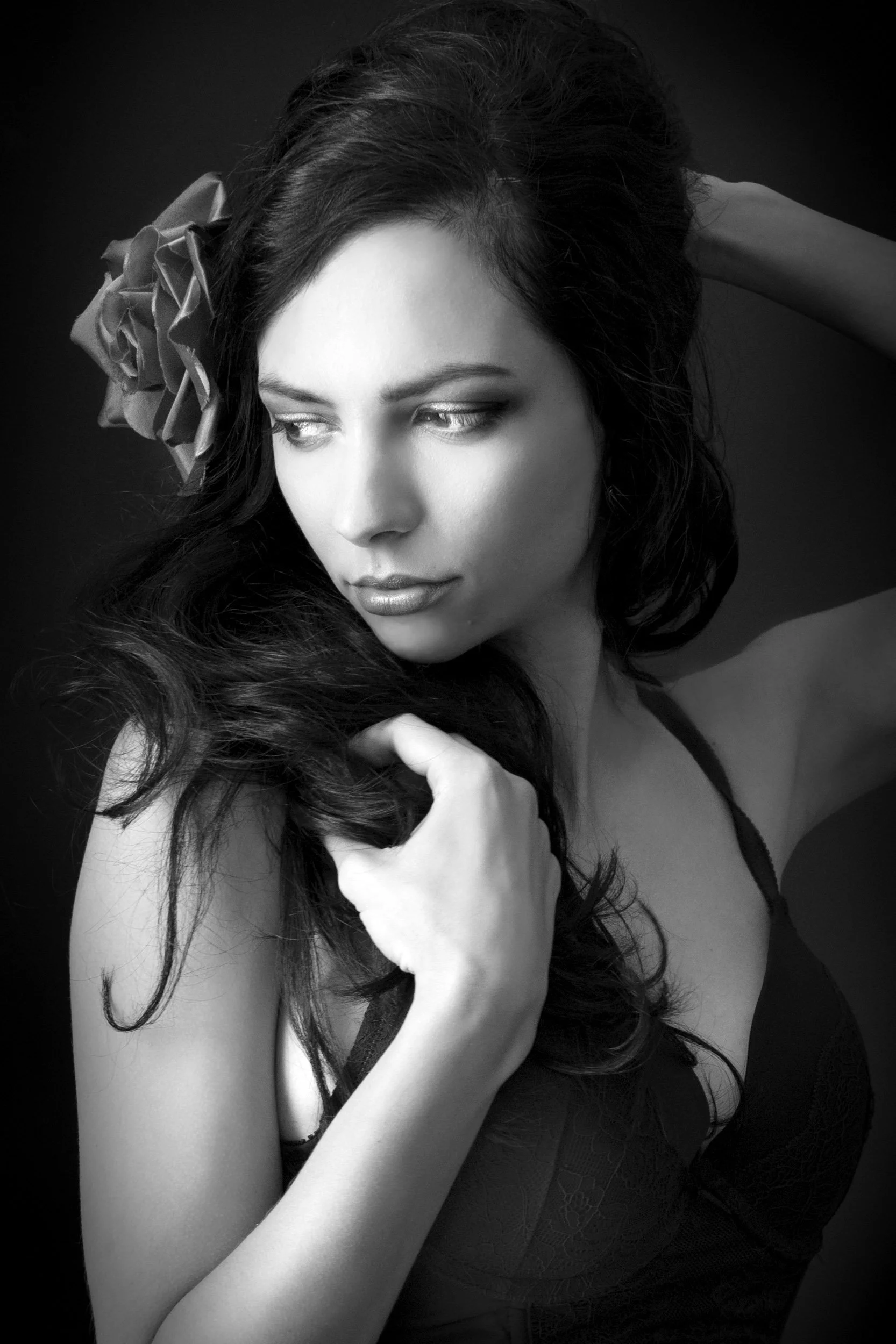 Black and white portrait of a woman with long dark hair and a large flower in her hair, looking to the side, with one hand touching her hair.